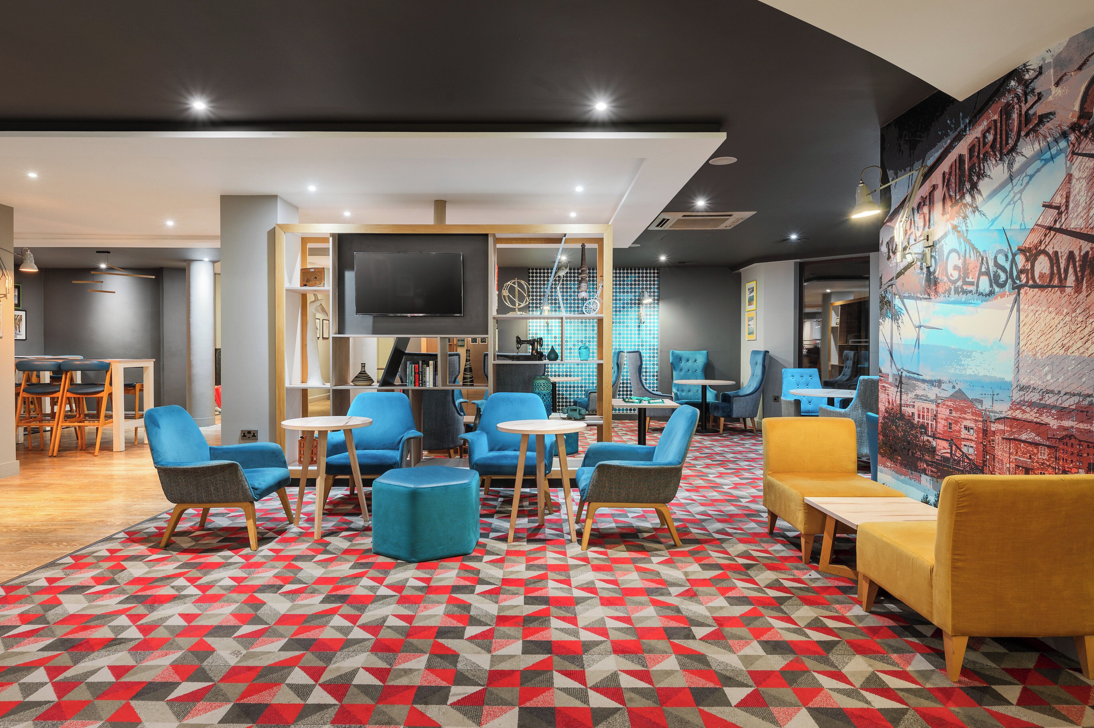 Ein moderner Loungebereich mit geometrisch gemustertem Teppich, blau-gelber Sitzgelegenheit, einem Flachbildfernseher und einem Wandgemälde von Glasgow.