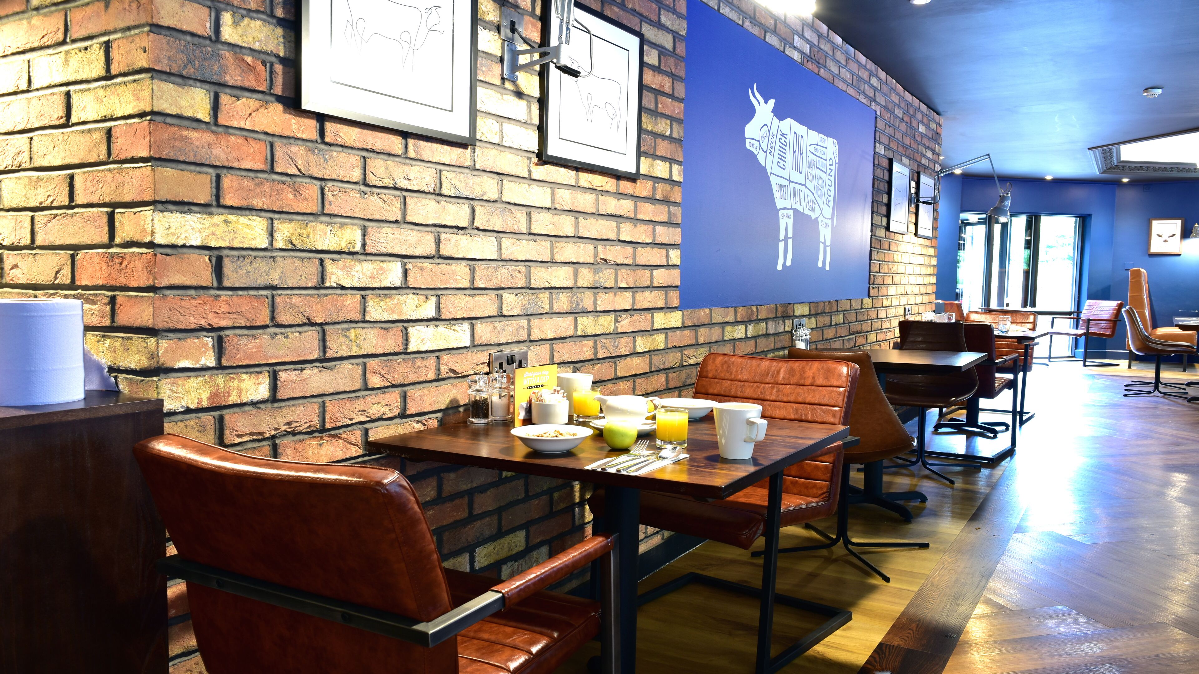 A restaurant with brick walls, framed artwork, and a blue wall with a cow silhouette. Tables are set with breakfast items.