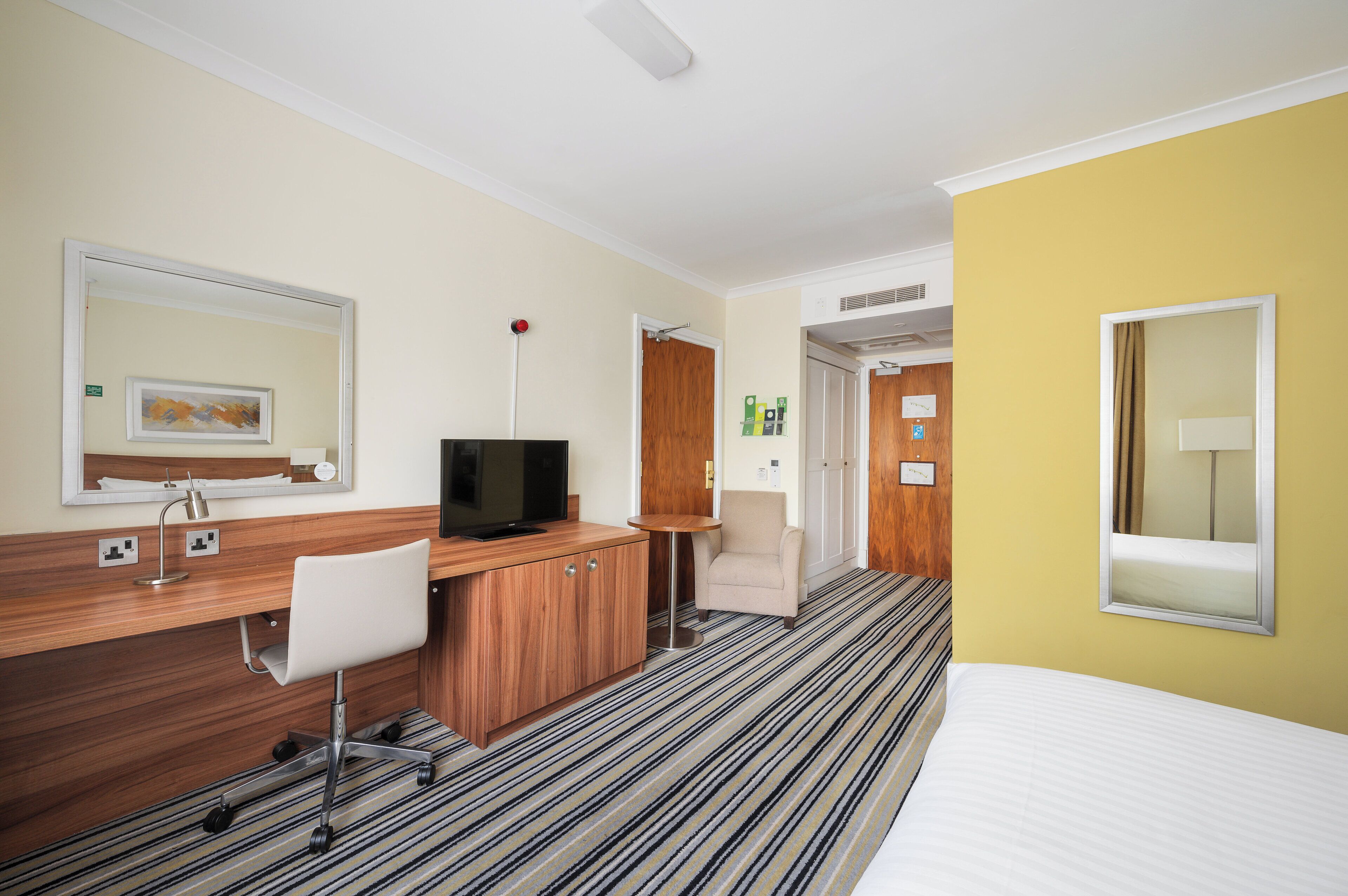 Habitación de hotel con escritorio de madera, televisor, una mesita redonda, una silla, cama y baño visible a través de una puerta abierta.