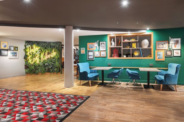 Interior moderno com parede verde, letreiro neon, piso de madeira e cadeiras azuis em volta de uma mesa.