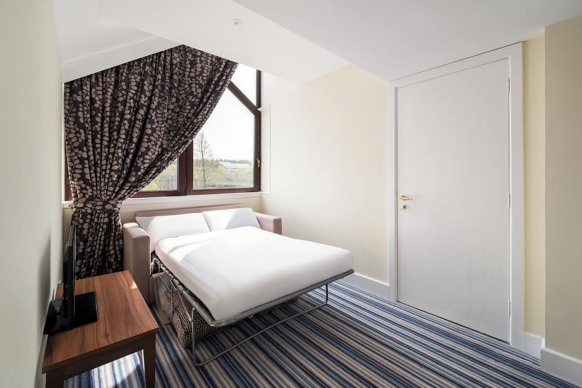 A hotel room with a bed, a desk, a window with curtains, and a door.