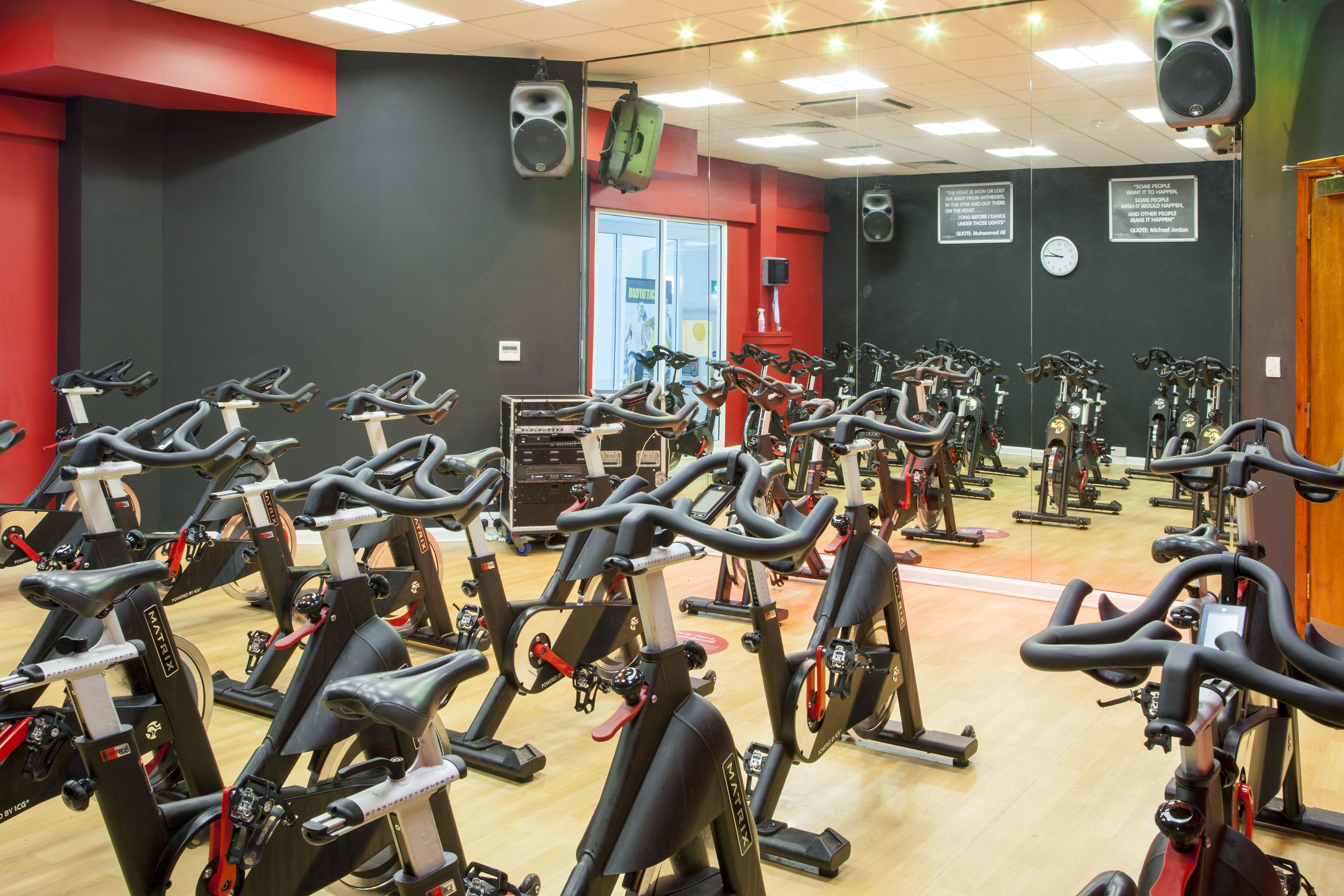 Un gimnasio con varias bicicletas estáticas dispuestas en filas, un reloj en la pared y un espejo que refleja las bicicletas.