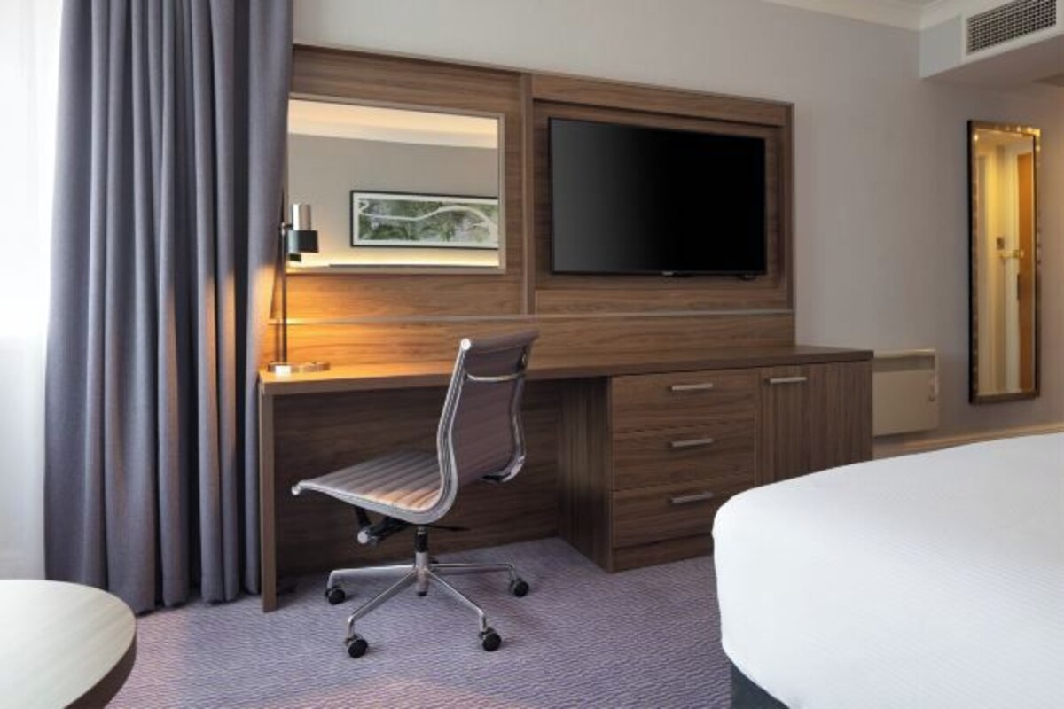 A hotel room with a wooden desk, a flat-screen TV, a large mirror, a bed, and a window with curtains.