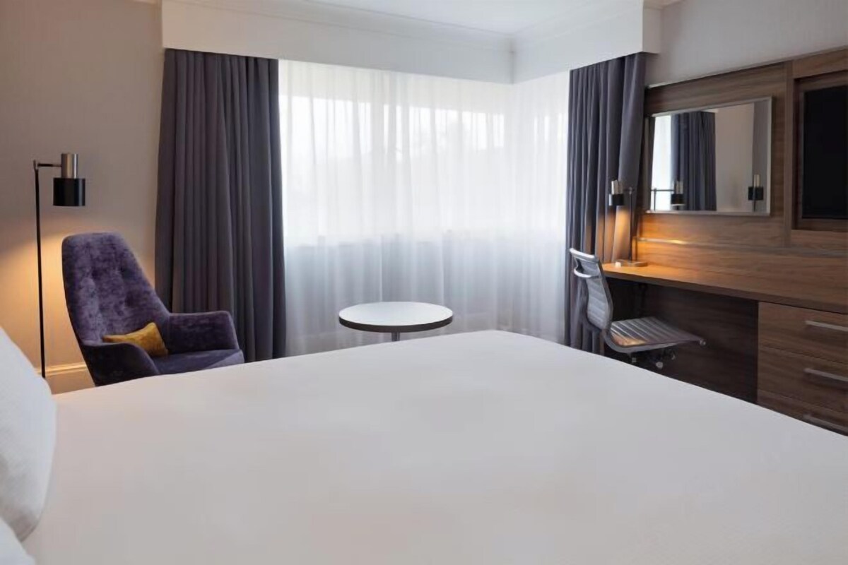 A hotel room with a large bed, a round table, a chair, a desk with a chair, a mirror, and a window with curtains.
