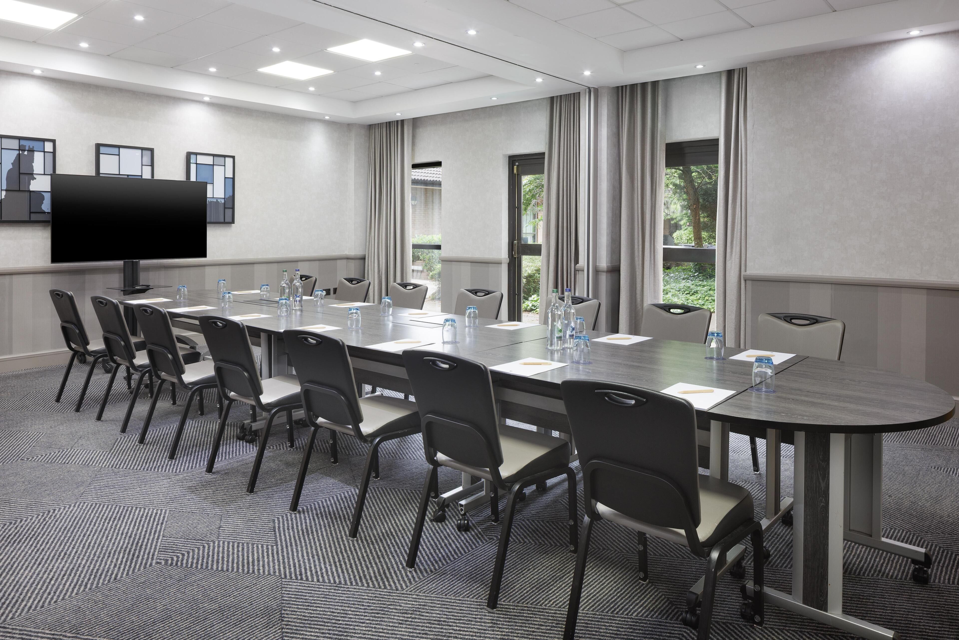 Un salón de conferencias con una larga mesa, sillas, un televisor de pantalla plana y ventanas con cortinas.