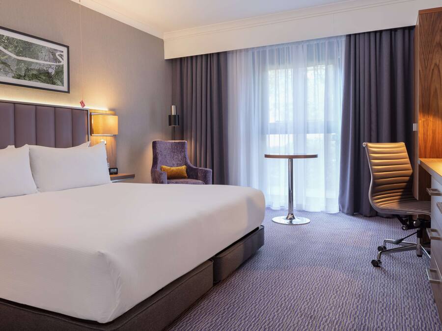 Ein Hotelzimmer mit einem großen Bett, einem Schreibtisch mit Stuhl, einem kleinen Tisch und einem Fenster mit Vorhängen.