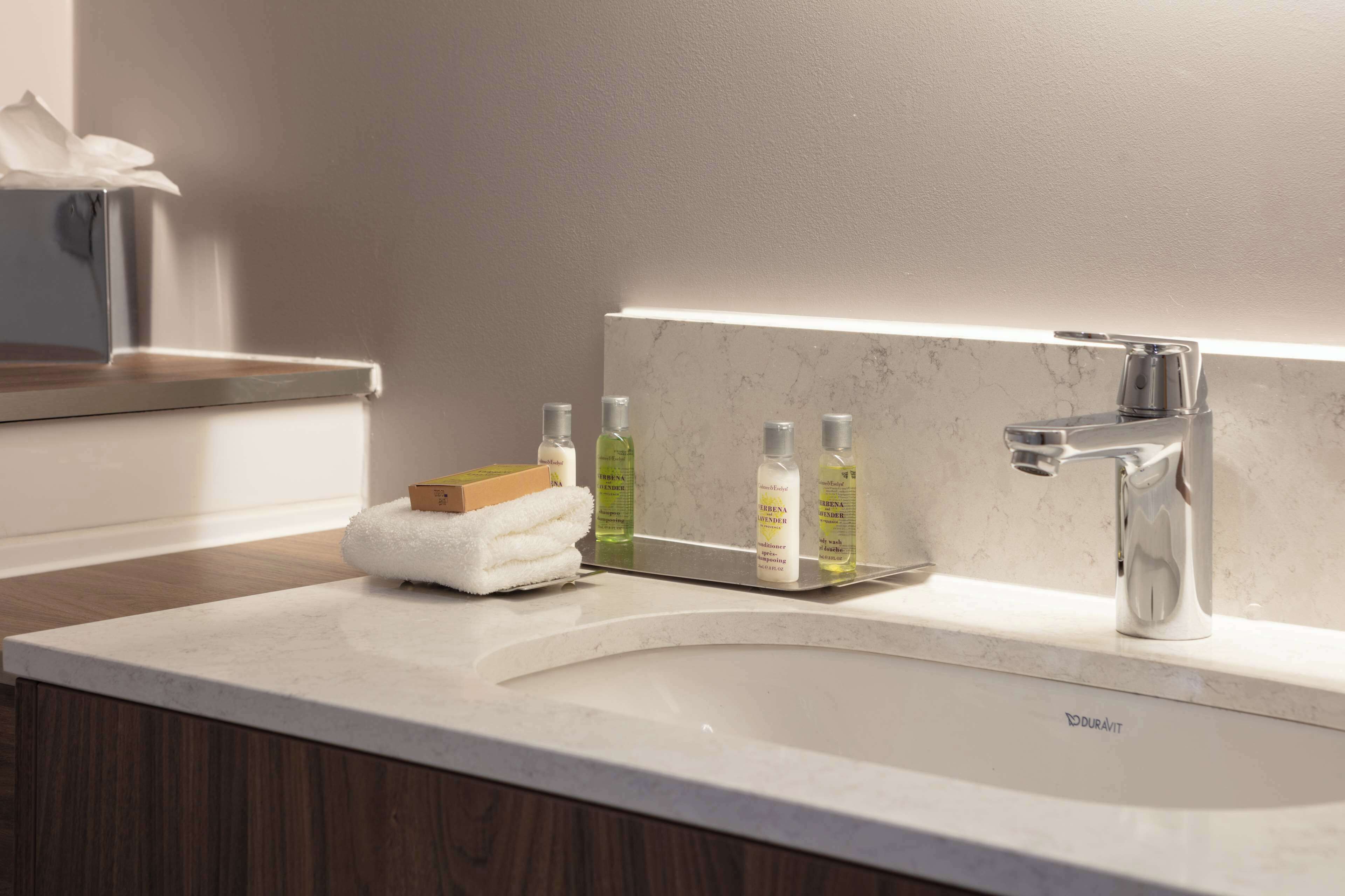 Ein modernes Badezimmer mit einem weißen Marmorwaschbecken, einem Chromarmaturen und einem Spiegel über dem Waschbecken. Auf dem Waschbecken liegen Pflegeprodukte und auf der Ablage ein Handtuch.