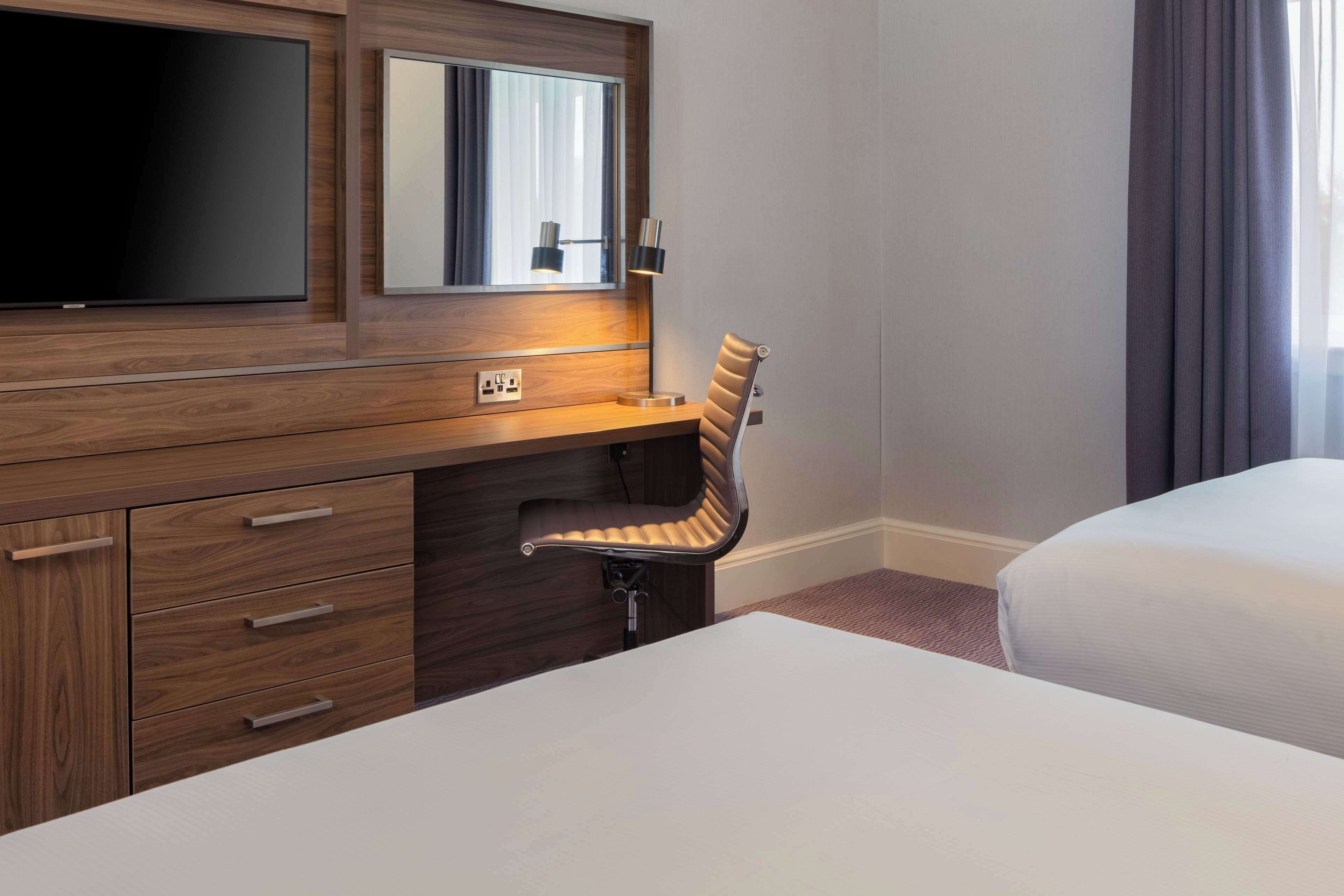 Una habitación de hotel con un escritorio de madera, un televisor de pantalla plana, un espejo, una lámpara de escritorio, una silla y una cama.