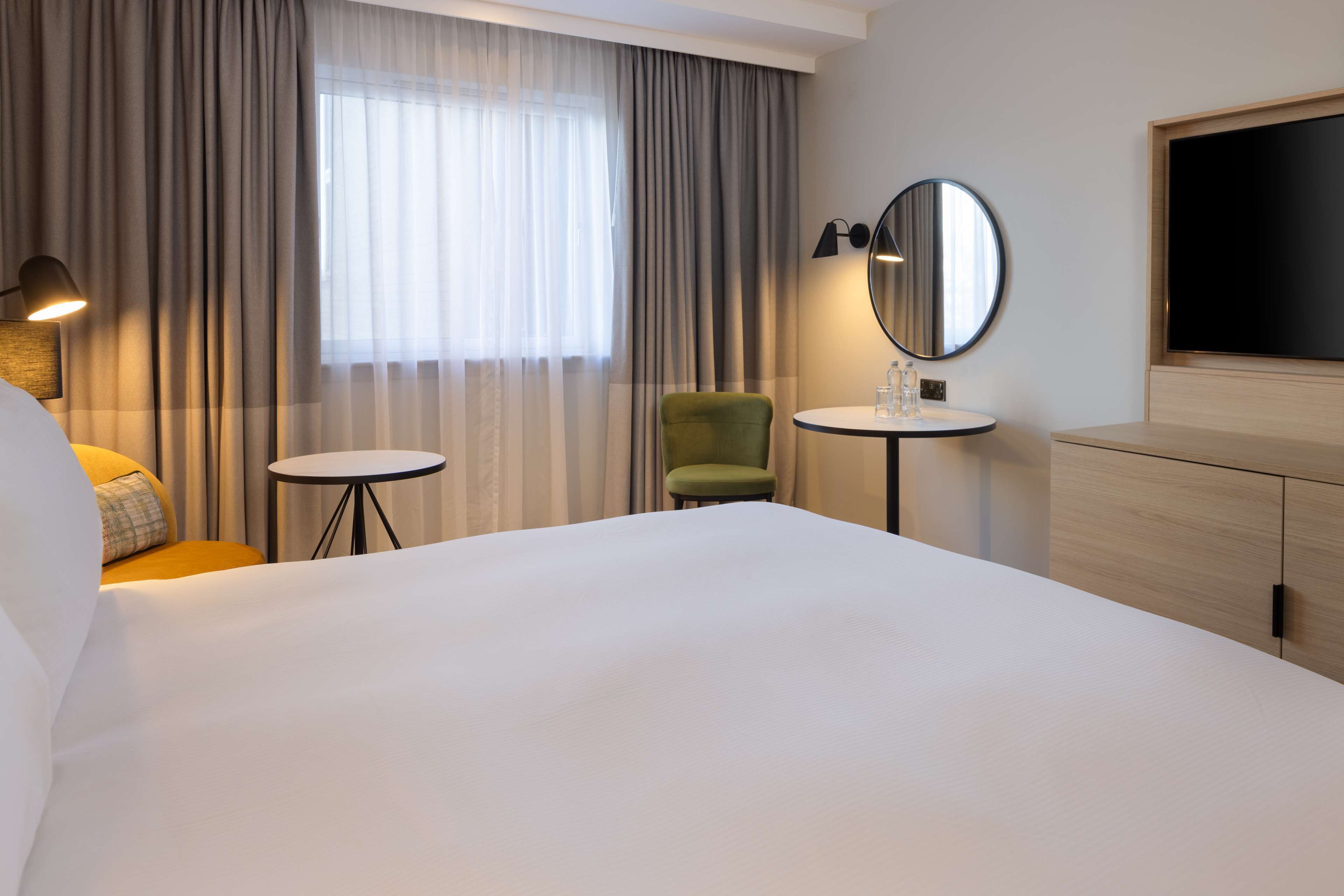 A hotel room with a large bed, a small round table, a green chair, a round mirror, and a TV mounted on the wall.