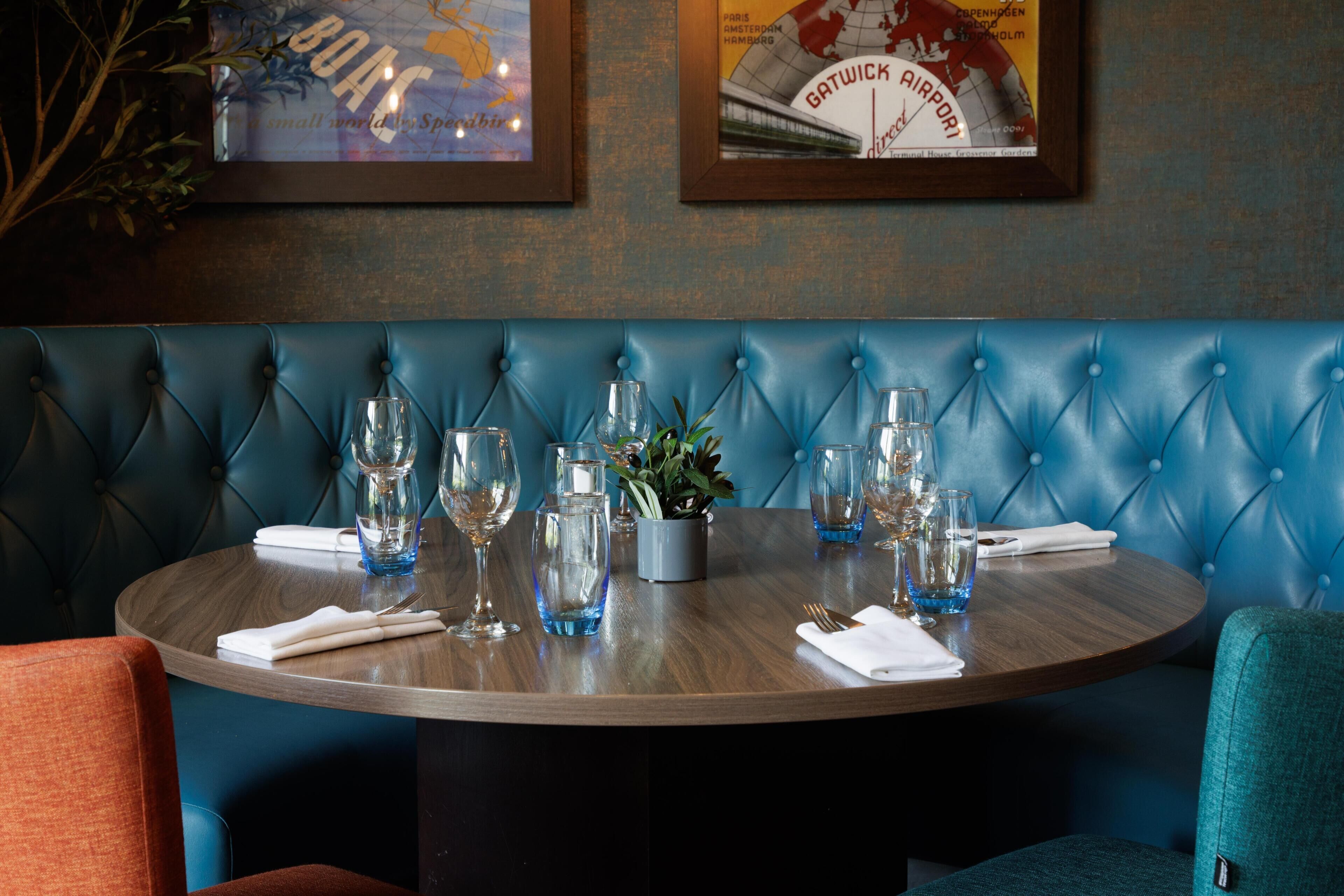 A round dining table with a blue upholstered bench, surrounded by empty wine glasses and water glasses, with a small potted plant on the table.