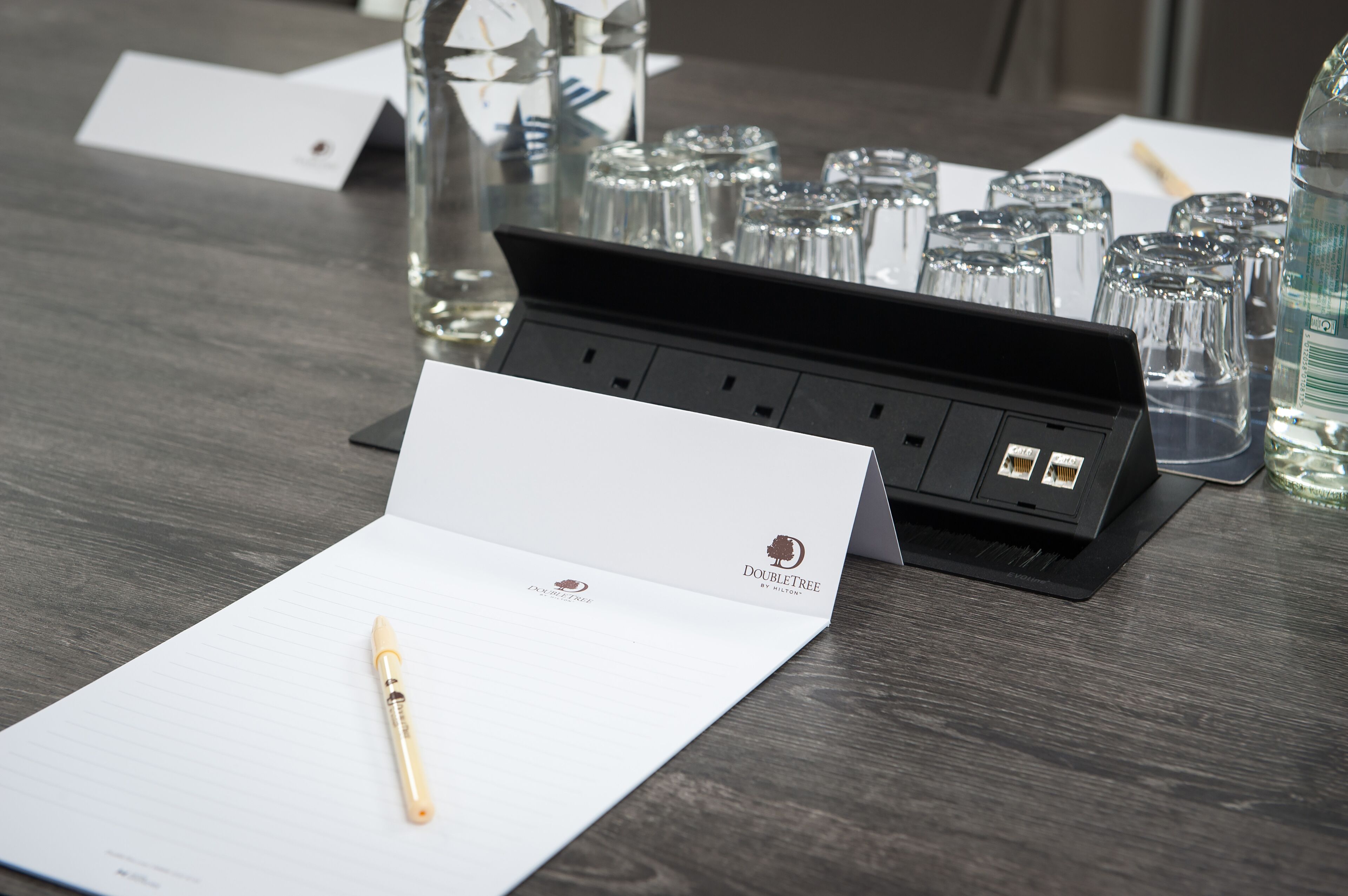 A hotel room with a wooden table, a pen, a notepad, a glass of water, and a glass set.