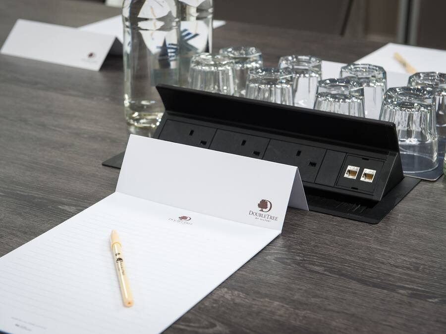 A hotel room with a wooden table, a pen, a notepad, a glass of water, and a glass set.