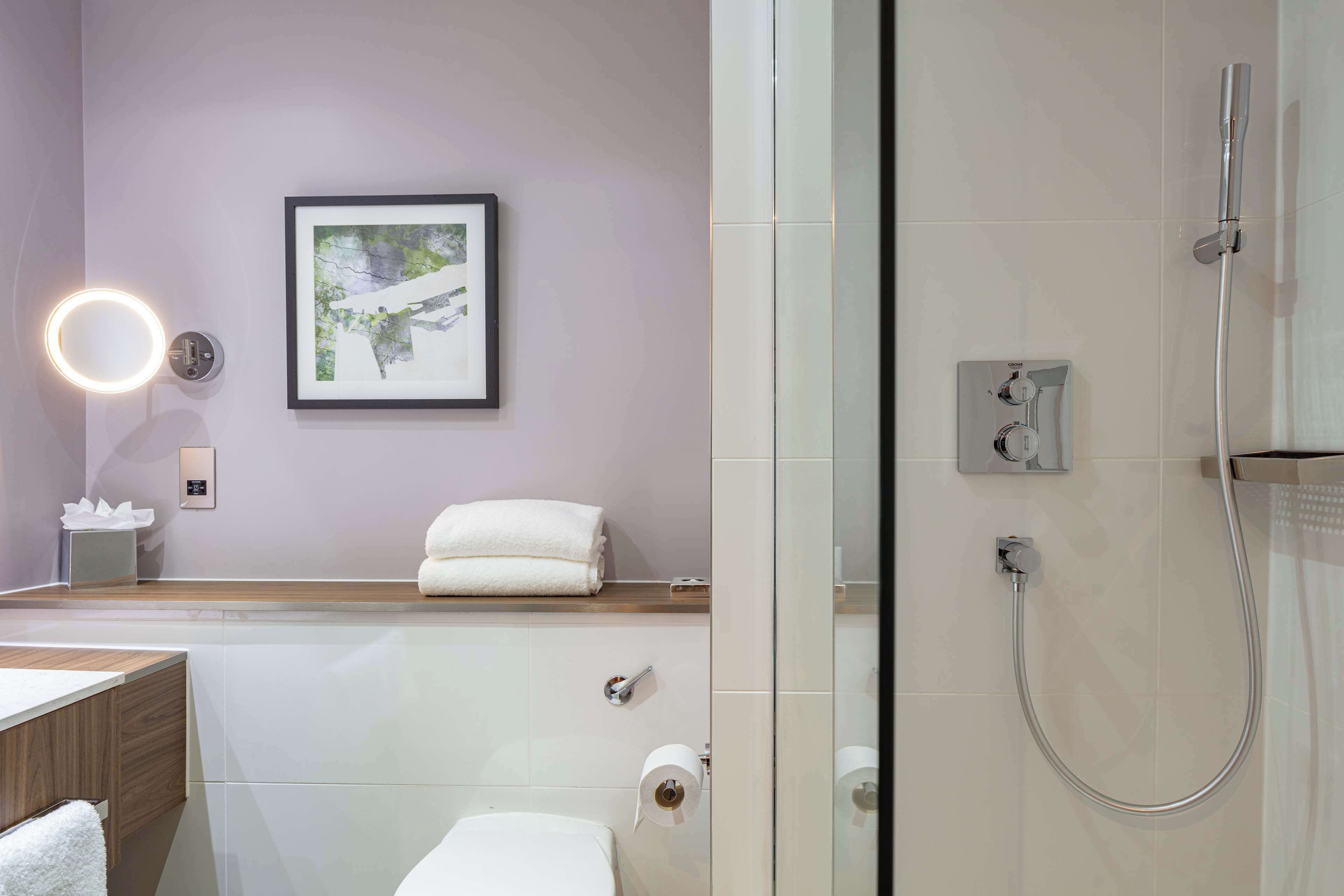 Une salle de bain moderne avec une douche à l’italienne en verre, une œuvre d’art encadrée au mur et un miroir rond avec un éclairage intégré.