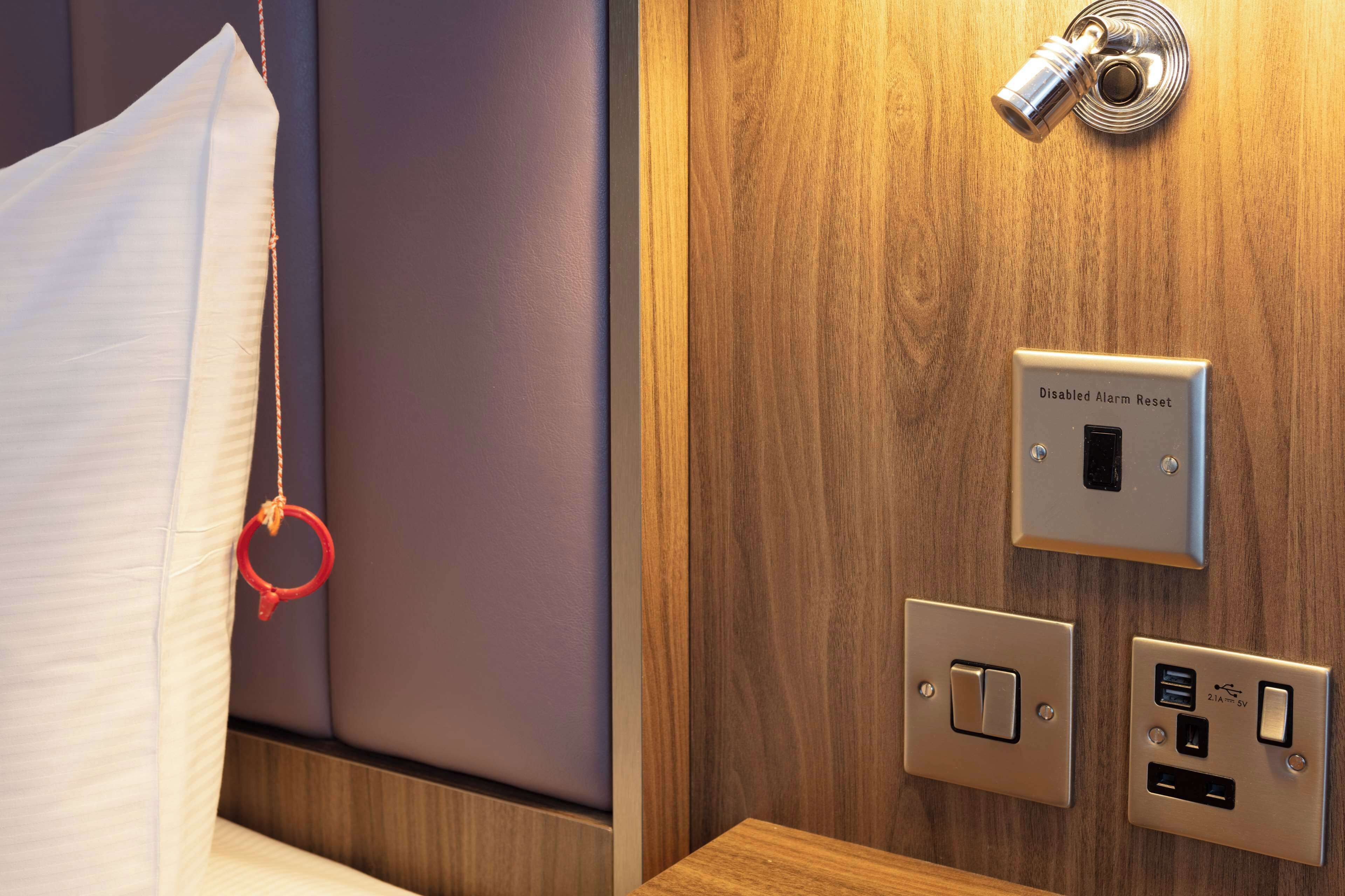 Habitación de hotel con cama, mesita de noche, un despertador en la pared y un panel de control con el botón 'Disabled Alarm Reset'.