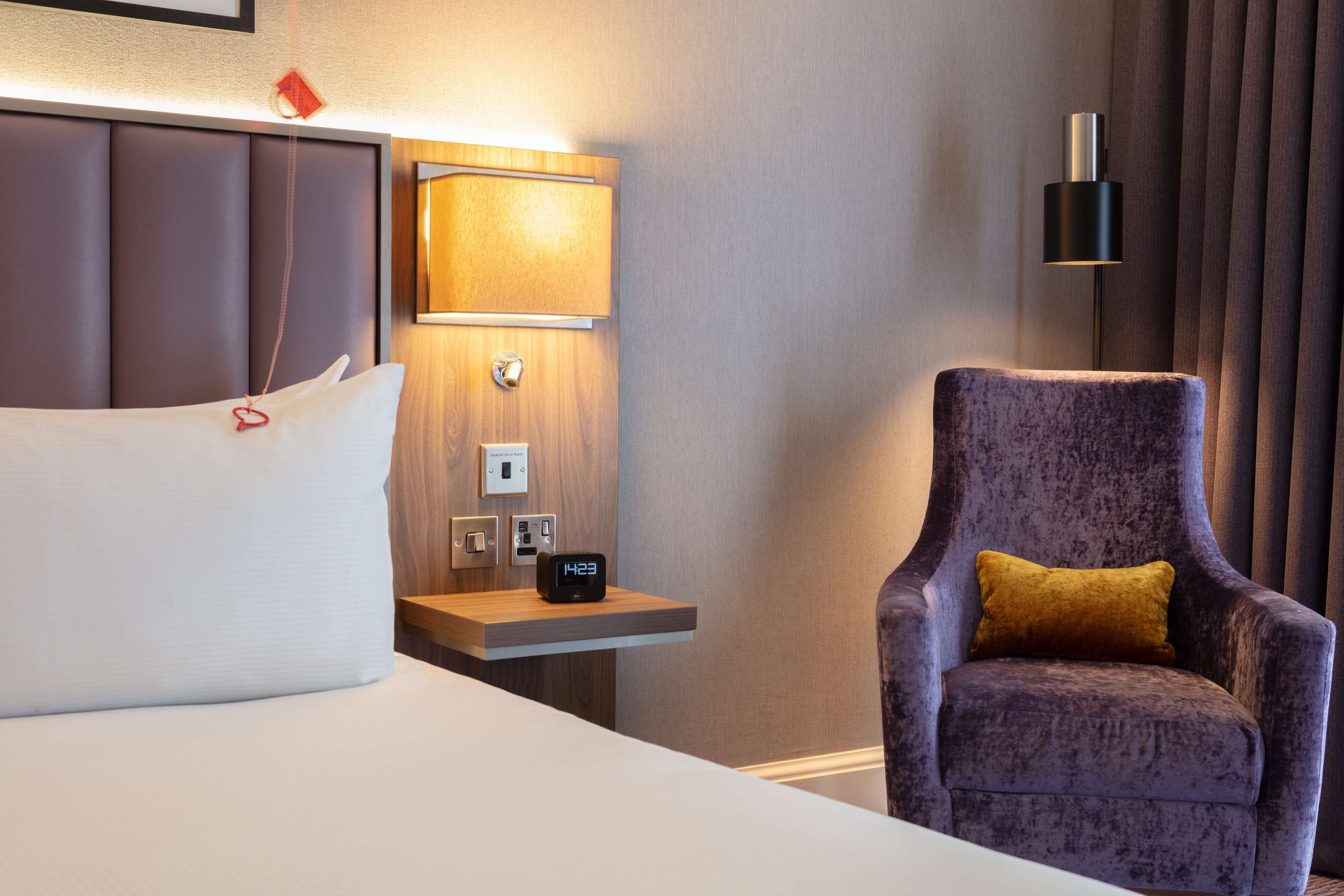 Una habitación de hotel con una cama, una mesita de noche con un reloj, una lámpara y una silla.