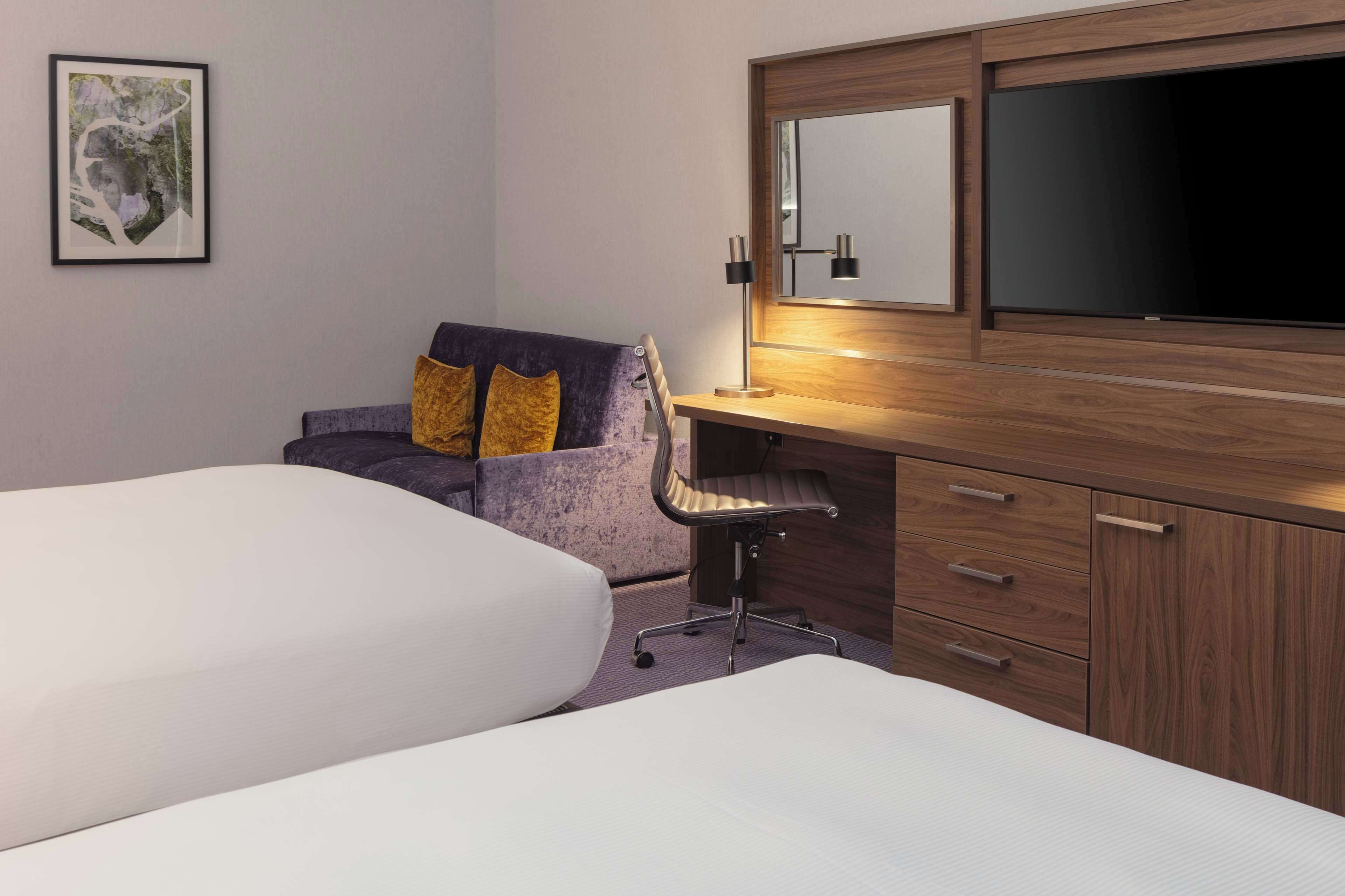 Une chambre d’hôtel avec deux lits, un bureau avec une chaise, un grand miroir et un tableau encadré au mur.