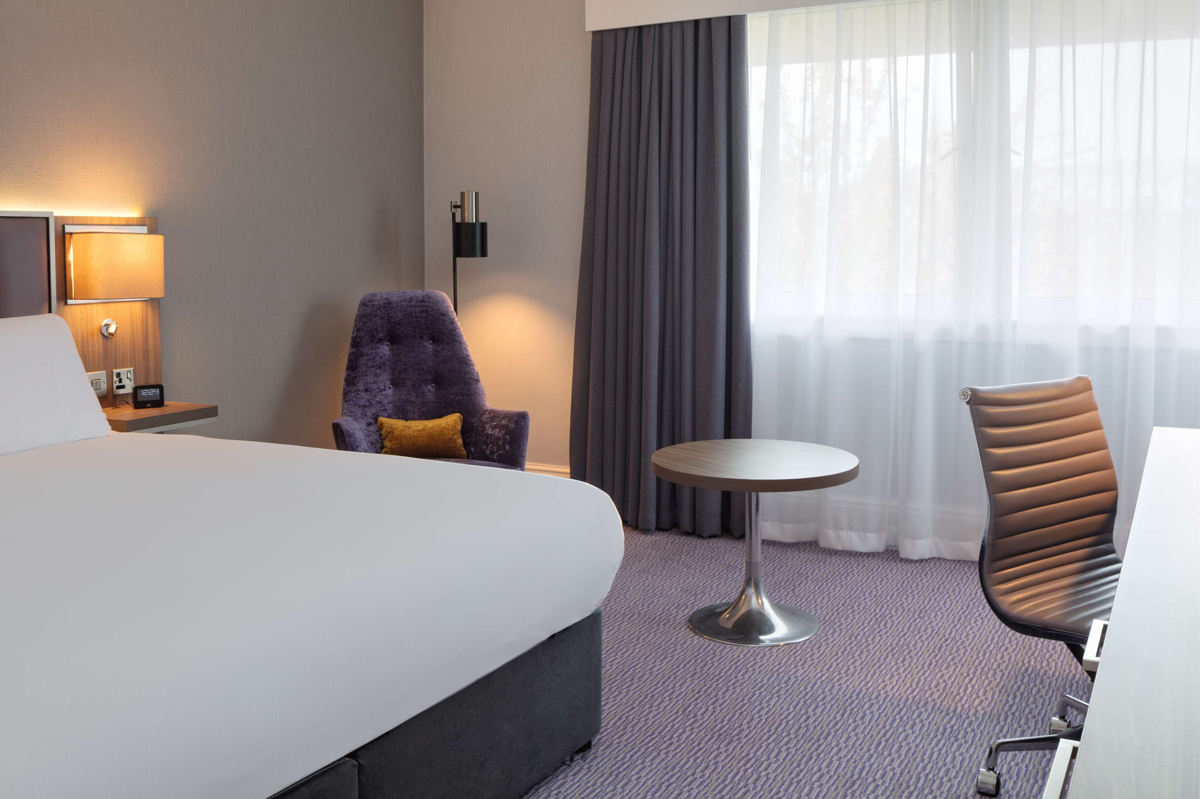 A hotel room with a large bed, a round table, two chairs, a lamp, and a window with curtains.