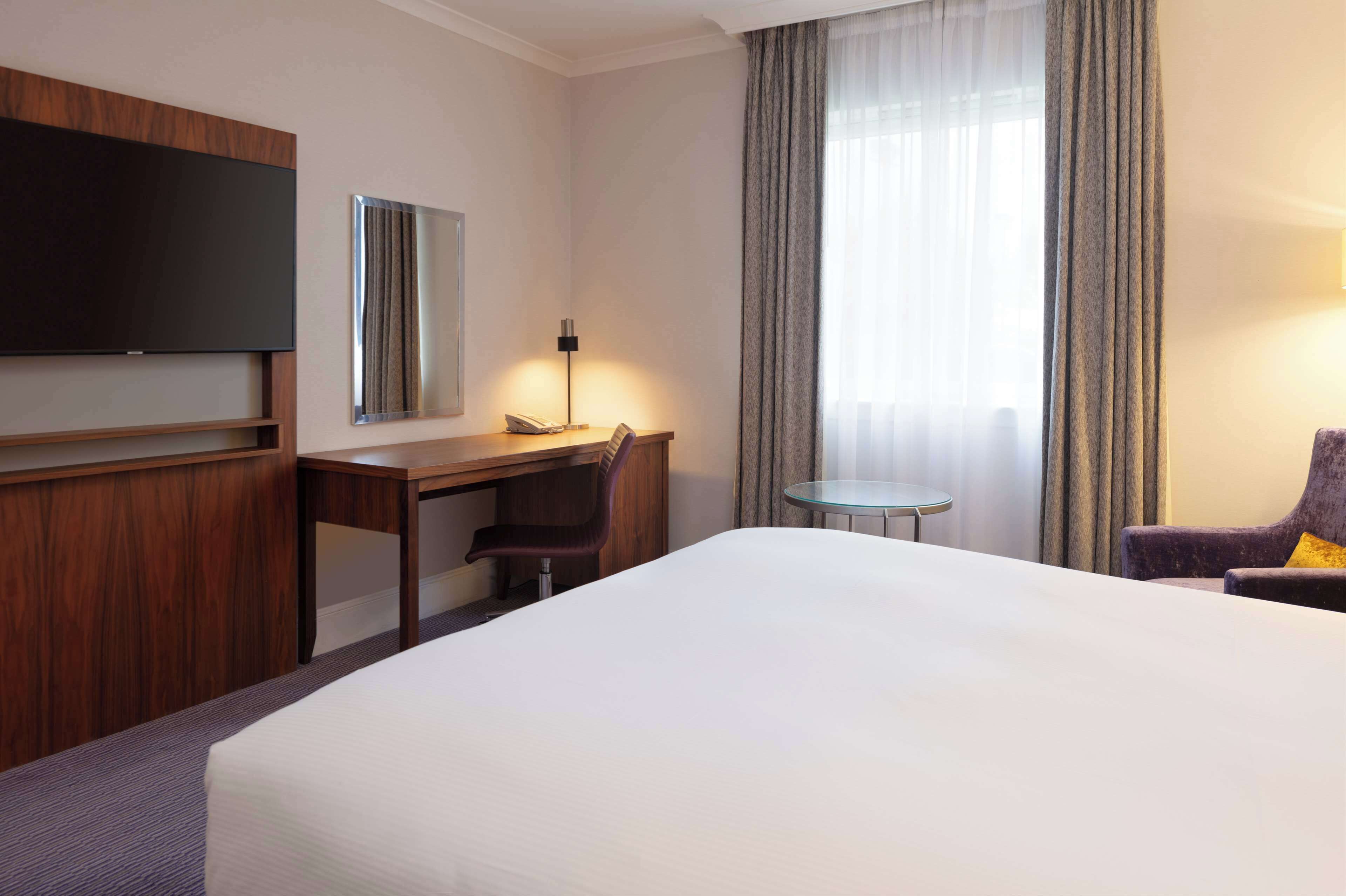 Habitación de hotel con una cama grande, un escritorio con silla, televisión, una mesita y una ventana con cortinas.