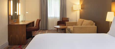 Ein Hotelzimmer mit einem großen Bett, einem Schreibtisch mit Stuhl, einem kleinen runden Tisch und zwei Sesseln.