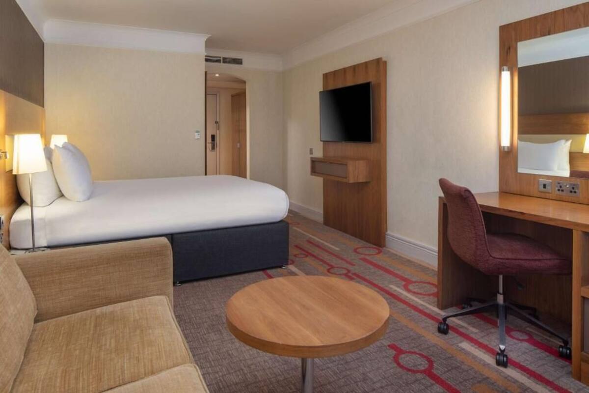 A hotel room with a bed, a desk, a chair, a TV, and a small round table.