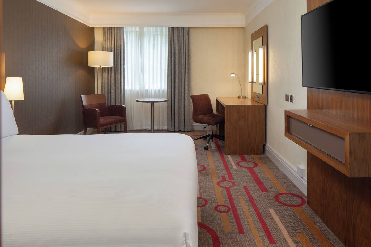 A hotel room with a large bed, a desk, two chairs, a TV, and a window with curtains.