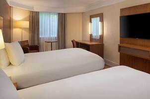 A hotel room with two beds, a desk, a chair, a lamp, a TV, and a window with curtains.