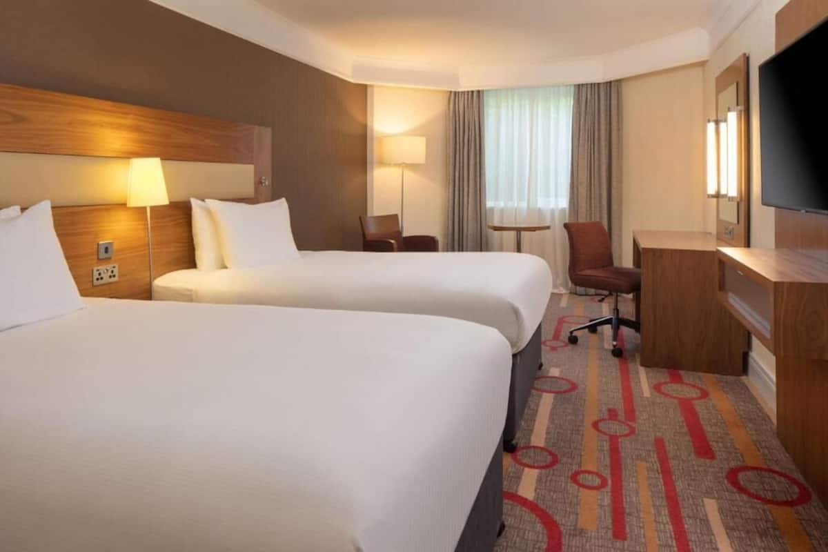 A hotel room with two beds, a desk, a chair, a TV, and a window with curtains.