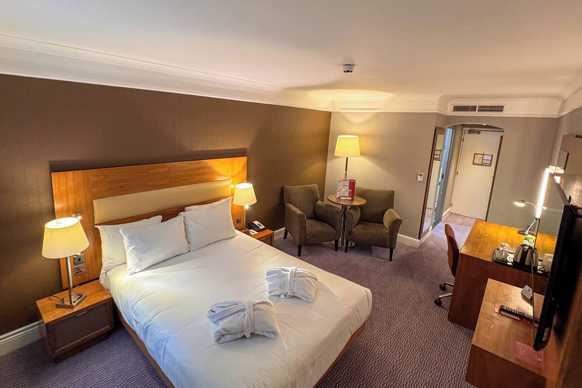 A hotel room with a large bed, two bedside lamps, a small table with a chair, a desk with a mirror, and a door.