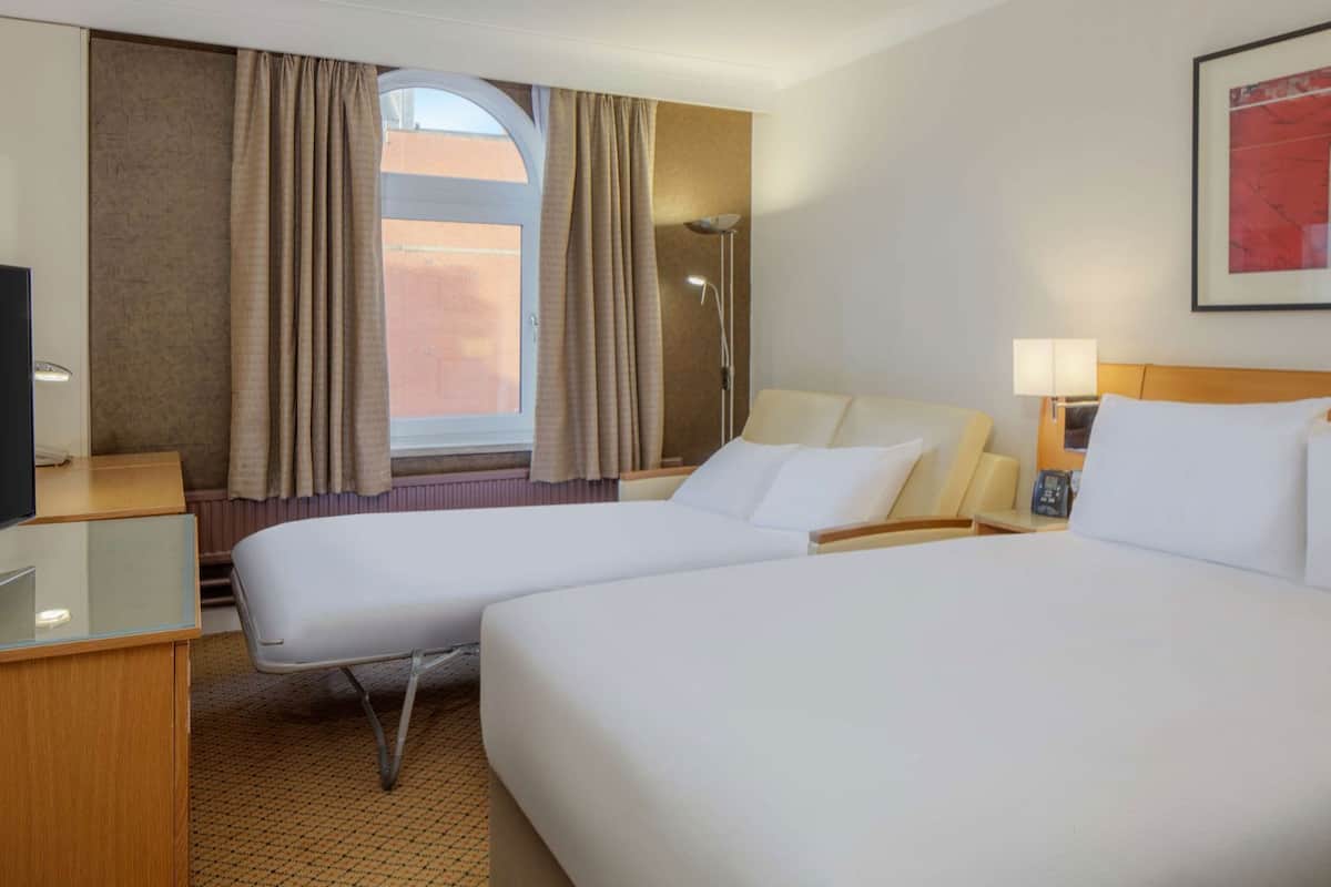 A hotel room with two beds, a desk, a chair, a lamp, and a window with curtains.