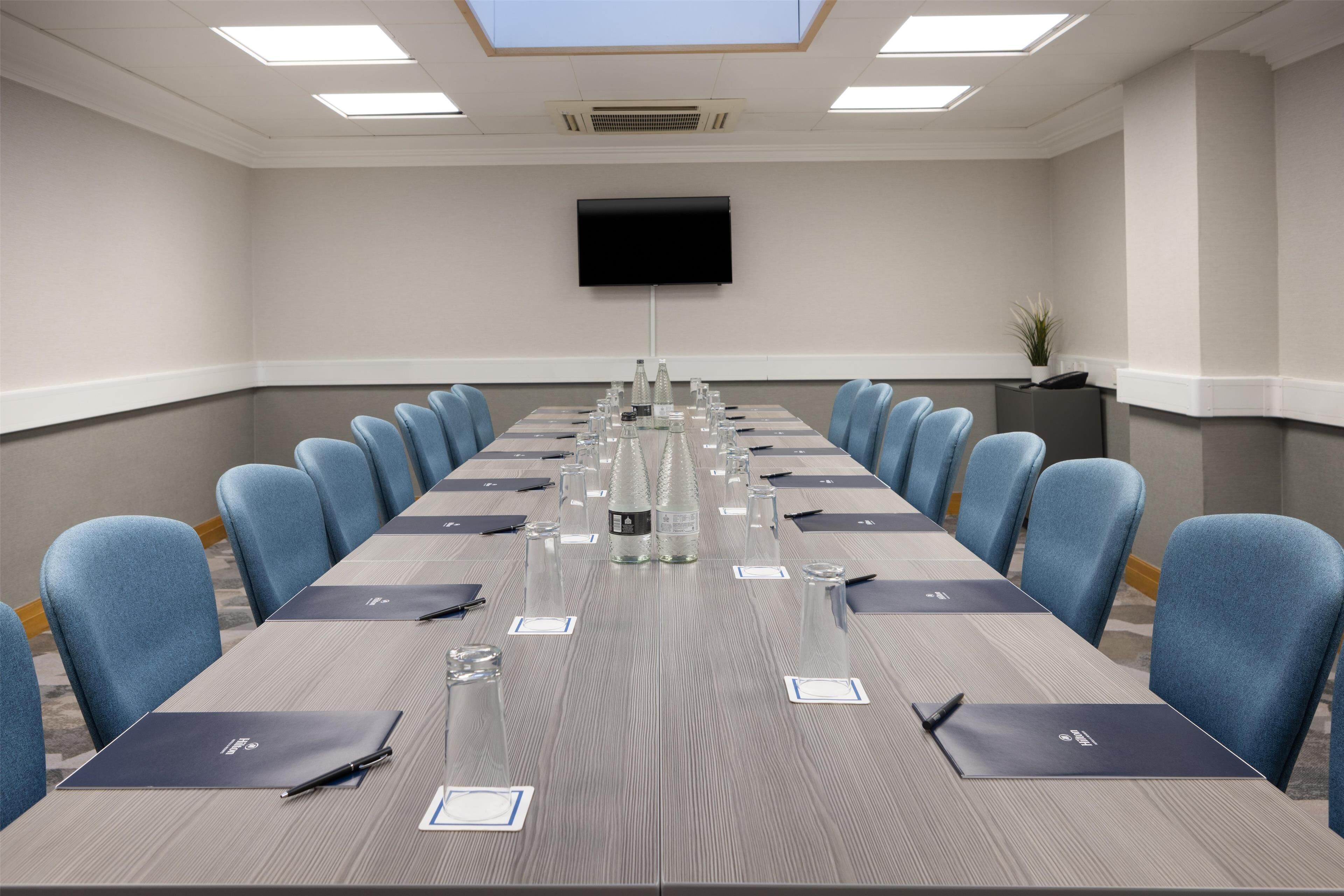 A conference room with a long table, blue chairs, name cards, water bottles, and a flat-screen TV on the wall.