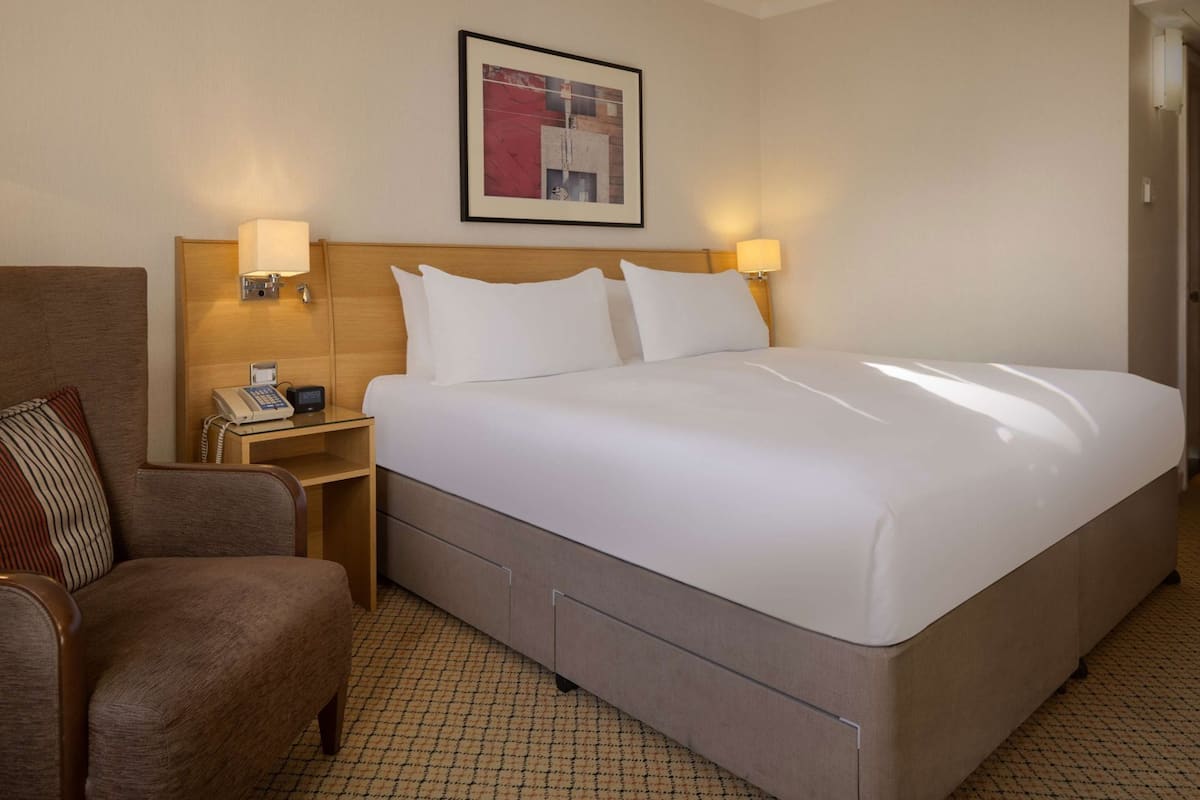 A hotel room with a bed, a chair, a bedside table with a phone, and a wall-mounted artwork.