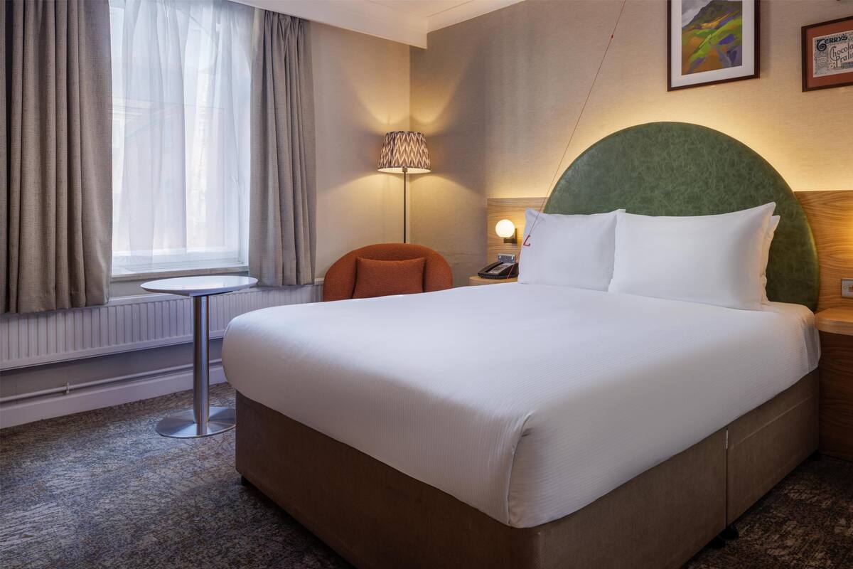 A hotel room with a large bed, a round green headboard, a small round table, a chair, a lamp, and a window with curtains.
