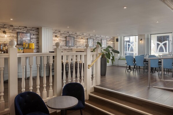 A modern hotel lobby with a wooden staircase, a reception desk, and a dining area with blue chairs.