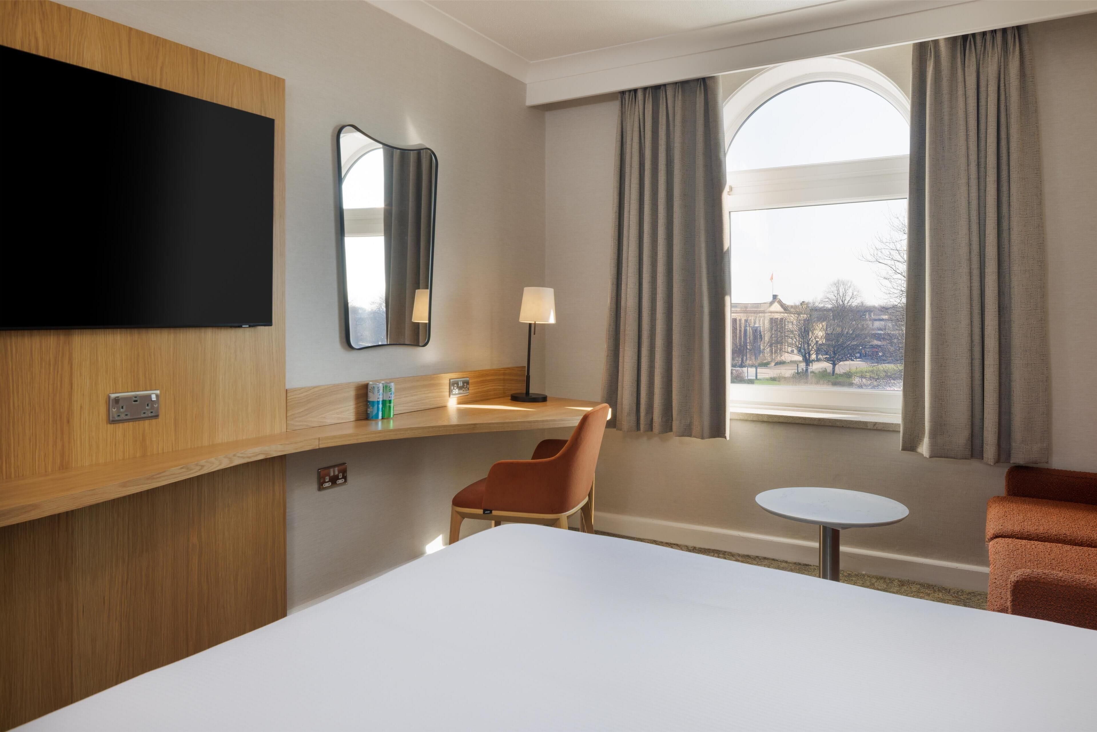 A hotel room with a bed, a TV, a desk with a lamp, a chair, a small table, and a window with curtains.