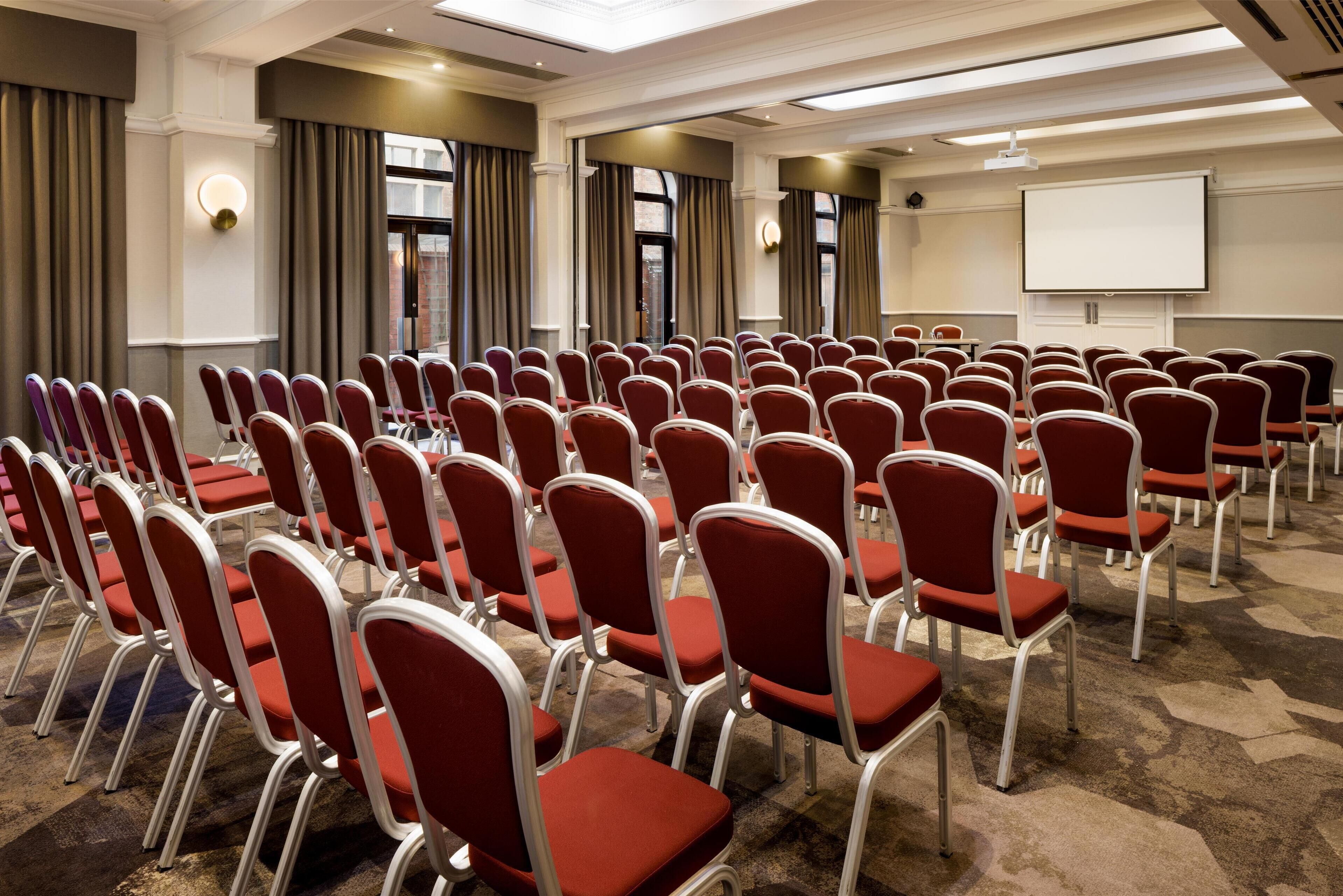 A conference room with rows of red chairs, a projector screen, and a carpeted floor.