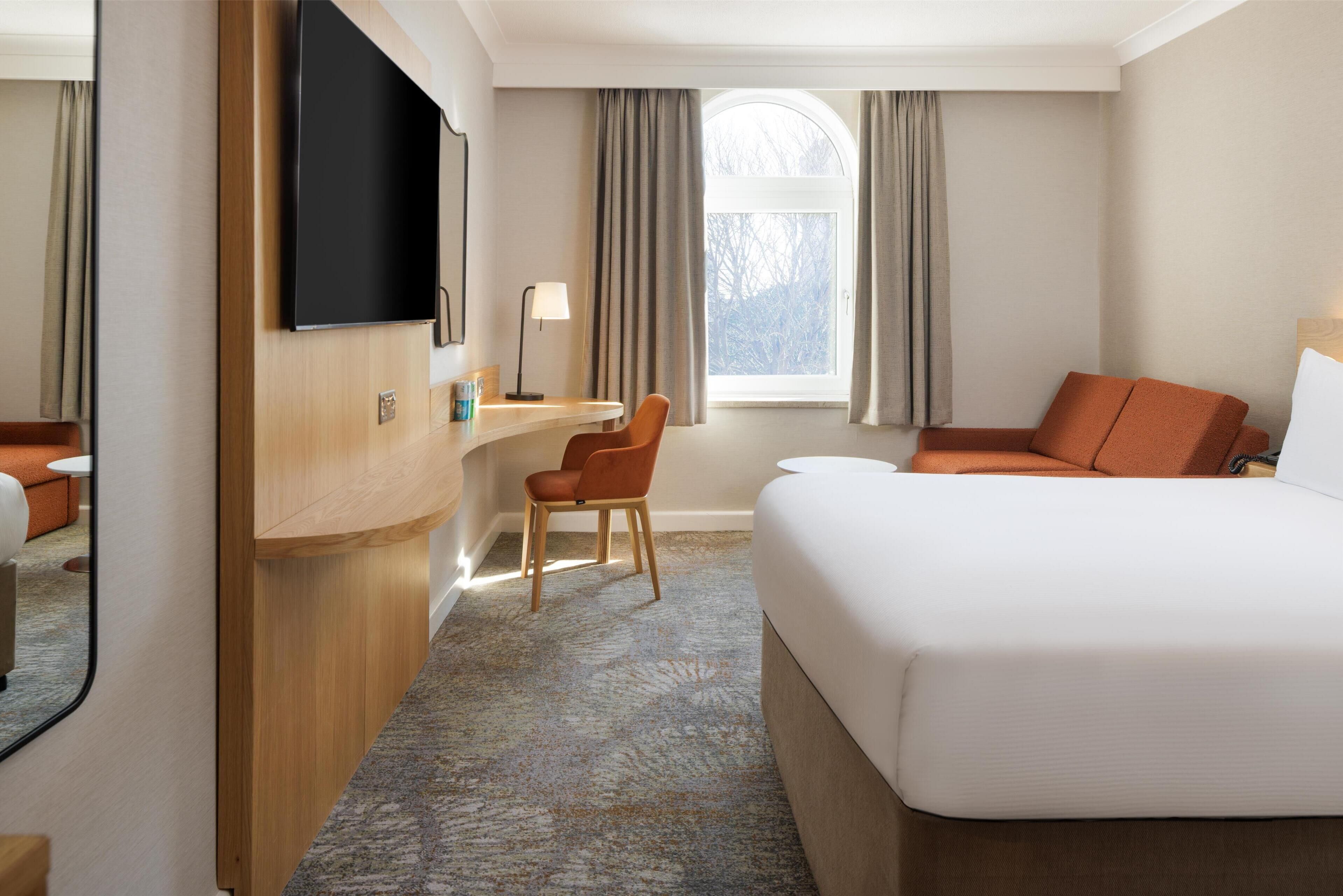 A hotel room with a large bed, a desk with a chair, a flat-screen TV mounted on the wall, and a window with curtains.