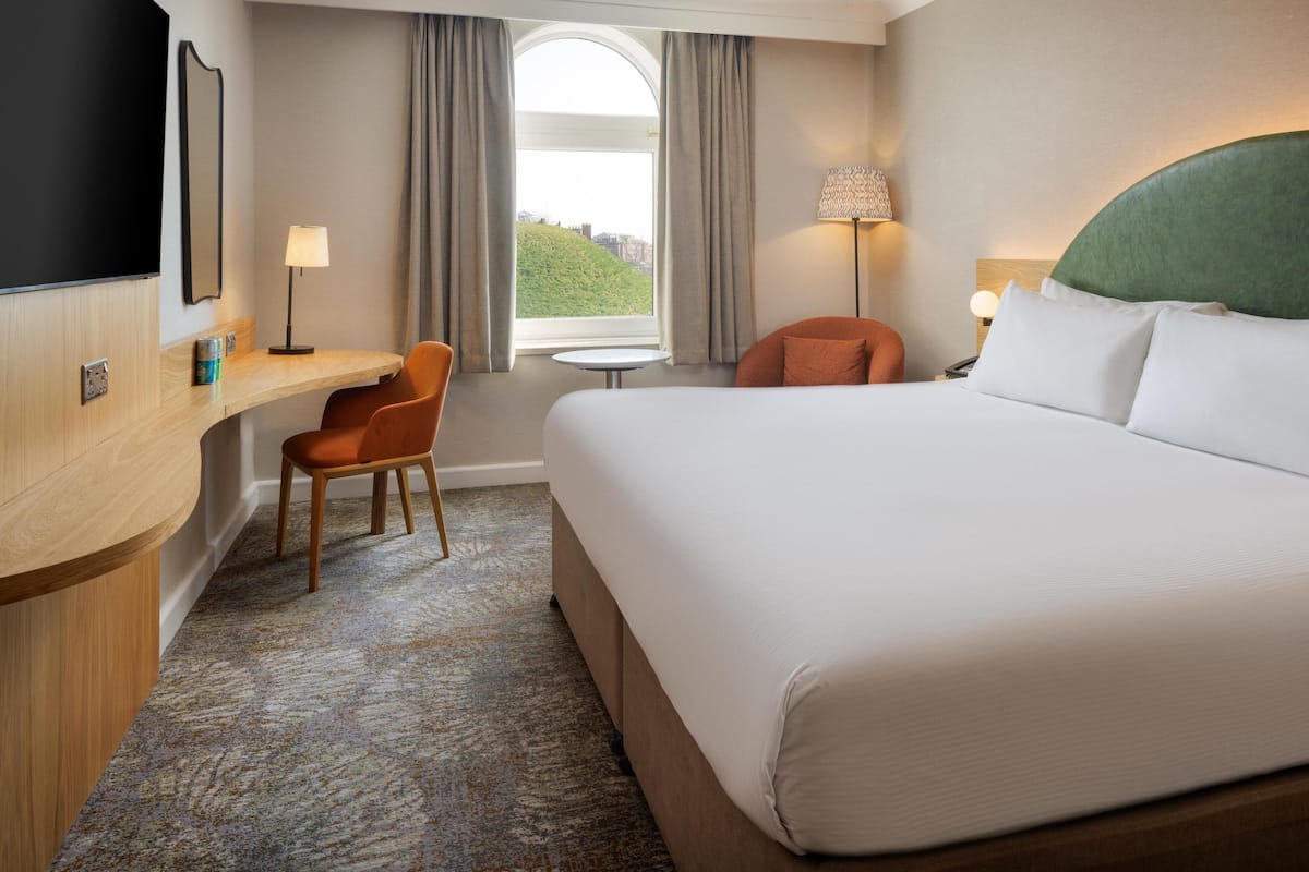 A hotel room with a large bed, a desk, a chair, a TV, and a window with a view of a hill.