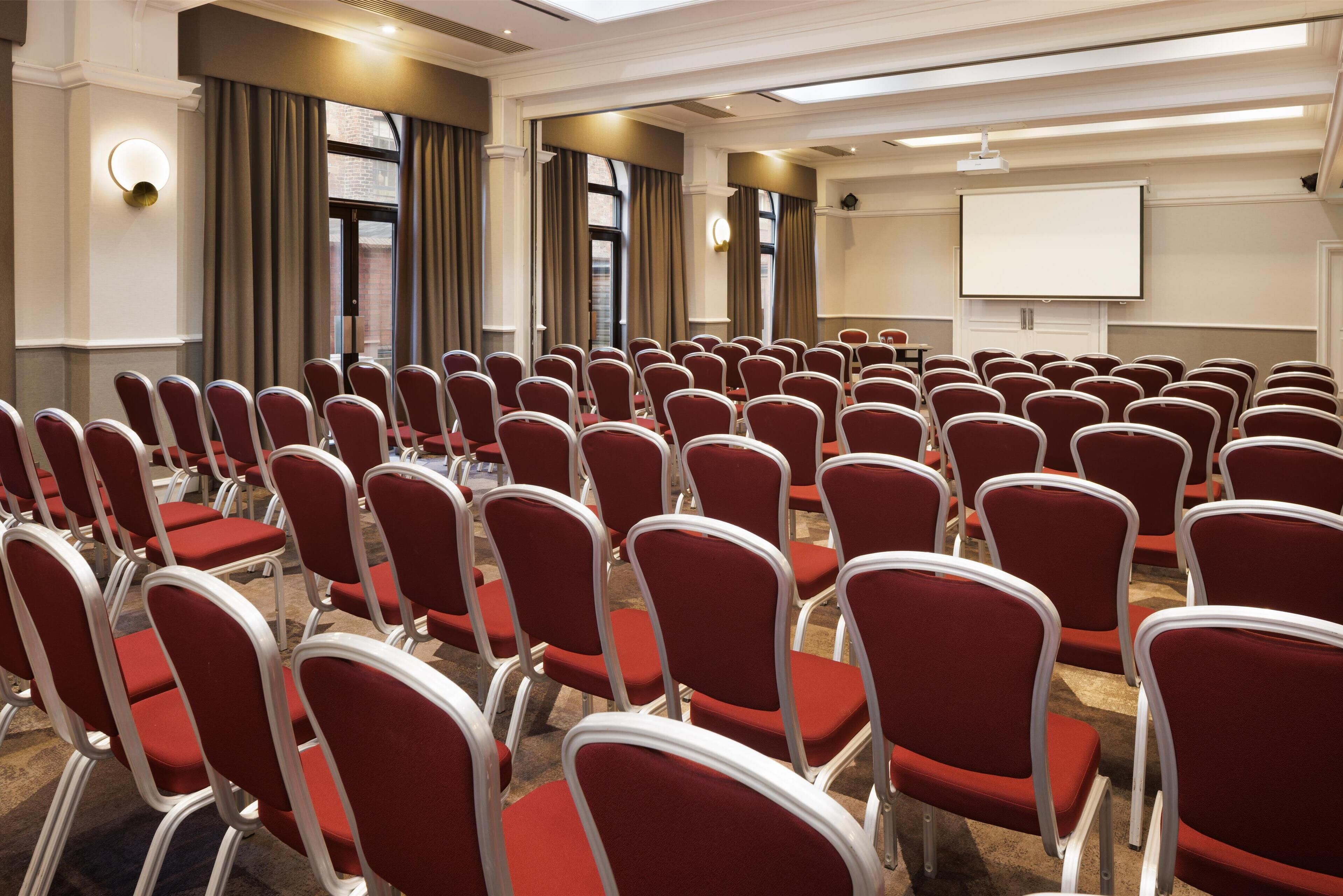 A conference room with rows of red chairs, a projector screen, and a large window with curtains.