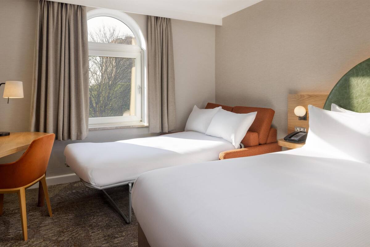 A hotel room with two beds, a desk, a chair, a lamp, and a view of trees through the window.