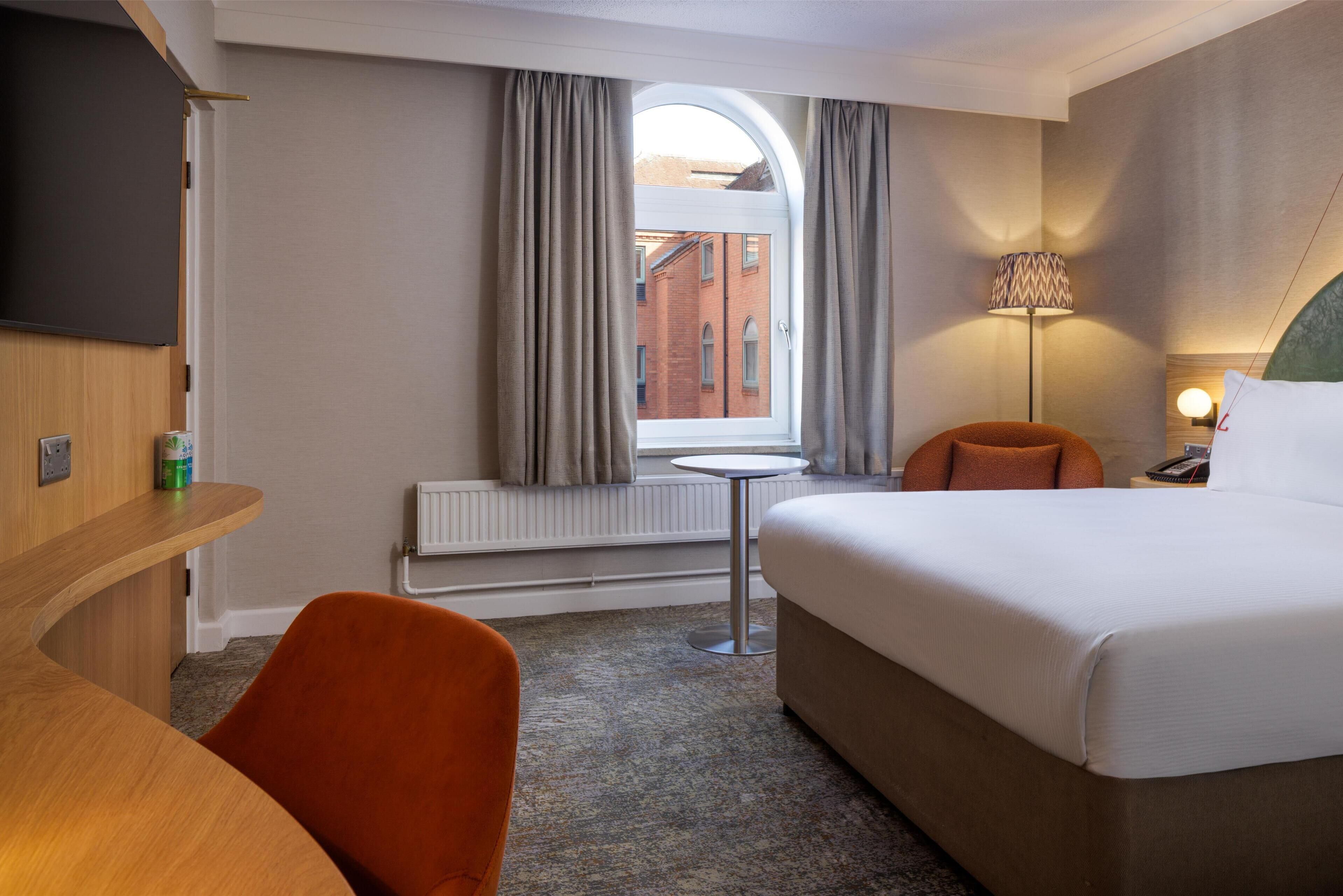 Une chambre d’hôtel avec un lit, un bureau, une chaise, une petite table, une lampe et une fenêtre avec des rideaux.