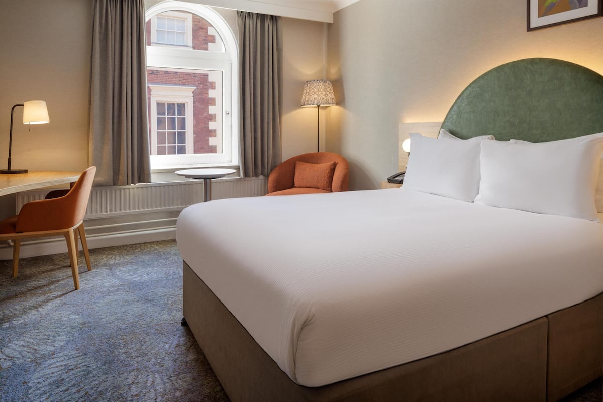 A hotel room with a large bed, a desk, a chair, a lamp, and a view of a building through a window.