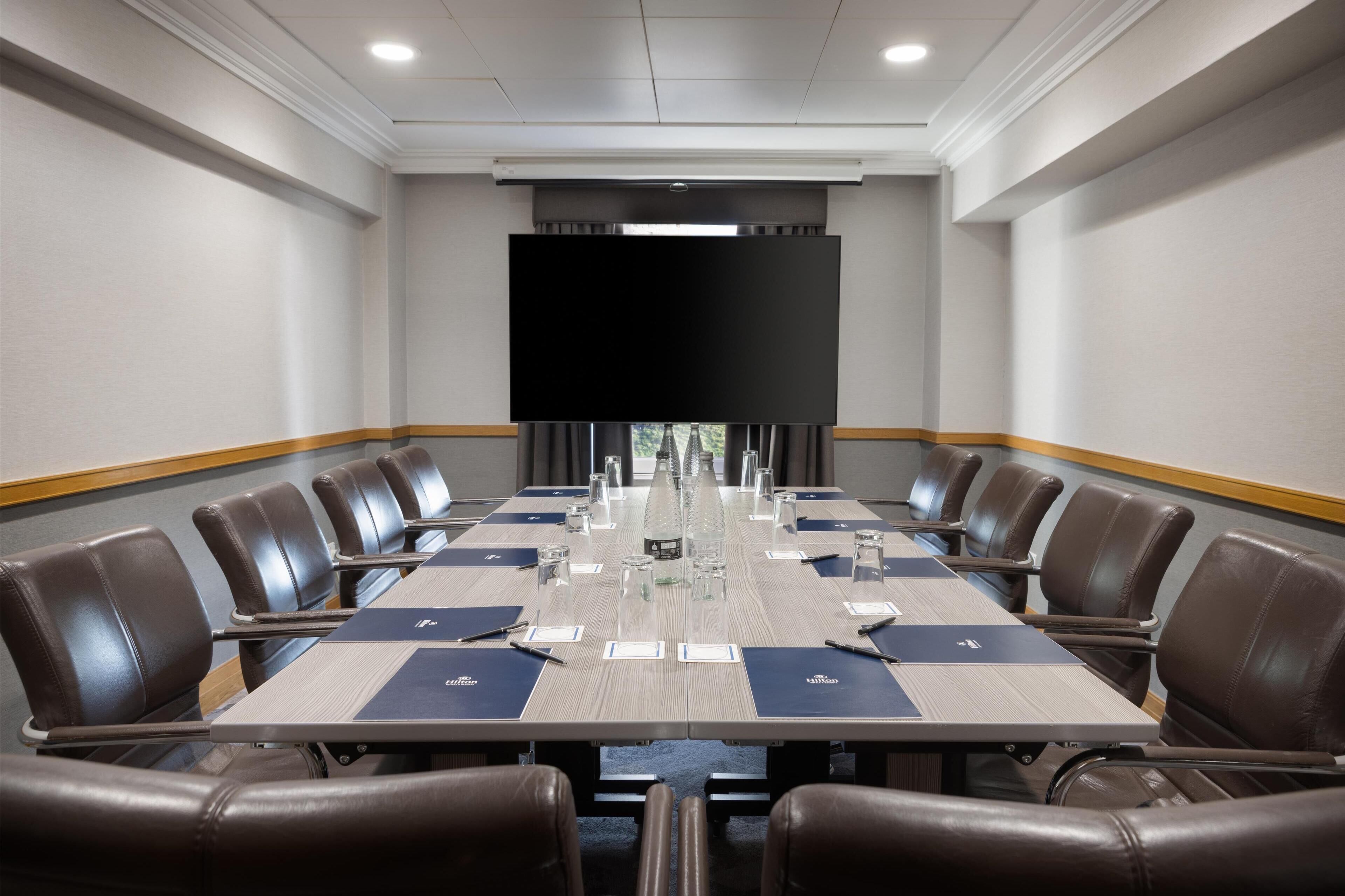 Ein Konferenzraum mit einem langen Tisch, braunen Ledersesseln, Namensschildern und einer großen Leinwand im Vordergrund.