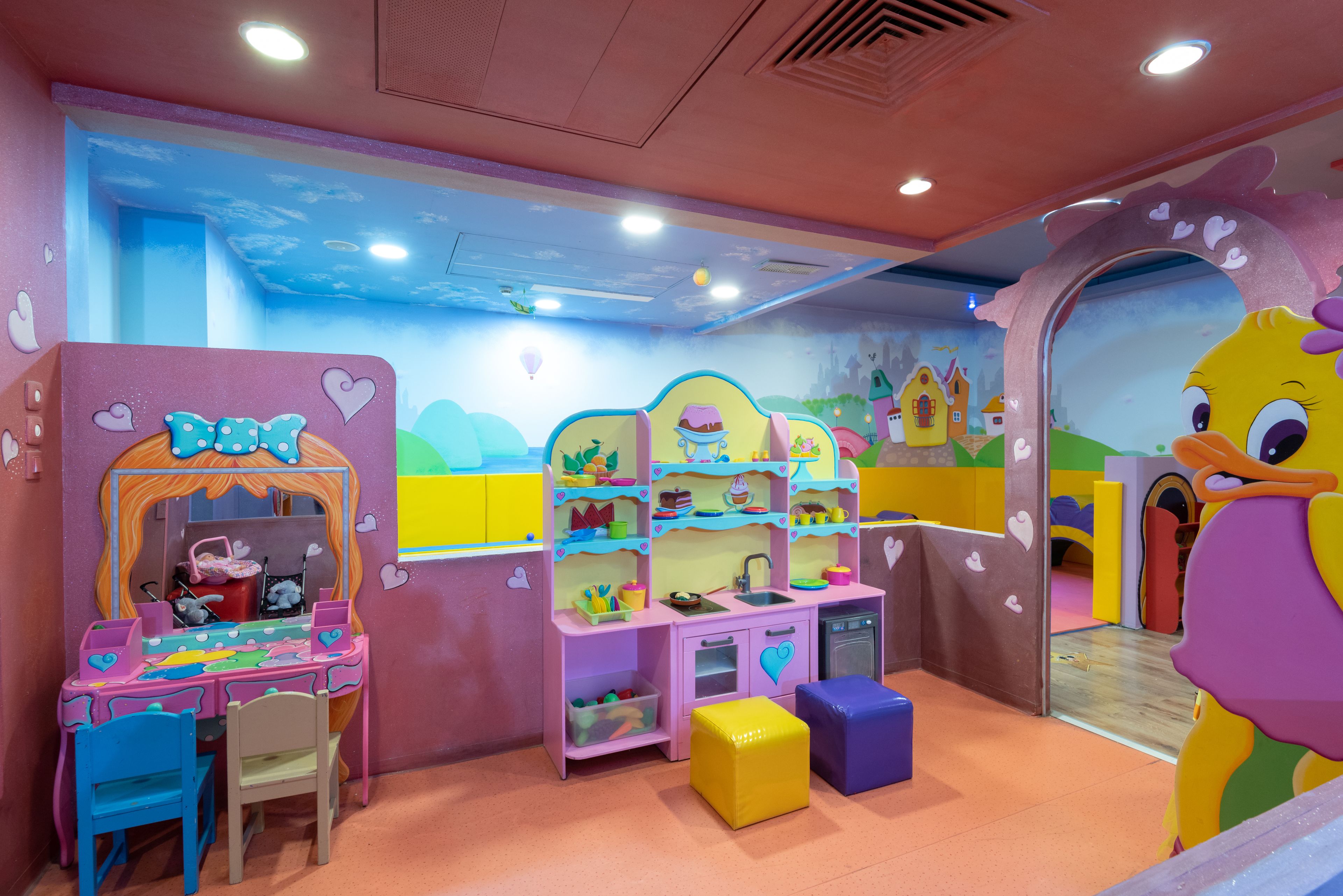 Un salón de juegos infantil con una decoración en tonos rosa y azul, que incluye una cocinita de juguete, un coche y un gran personaje de dibujos animados.