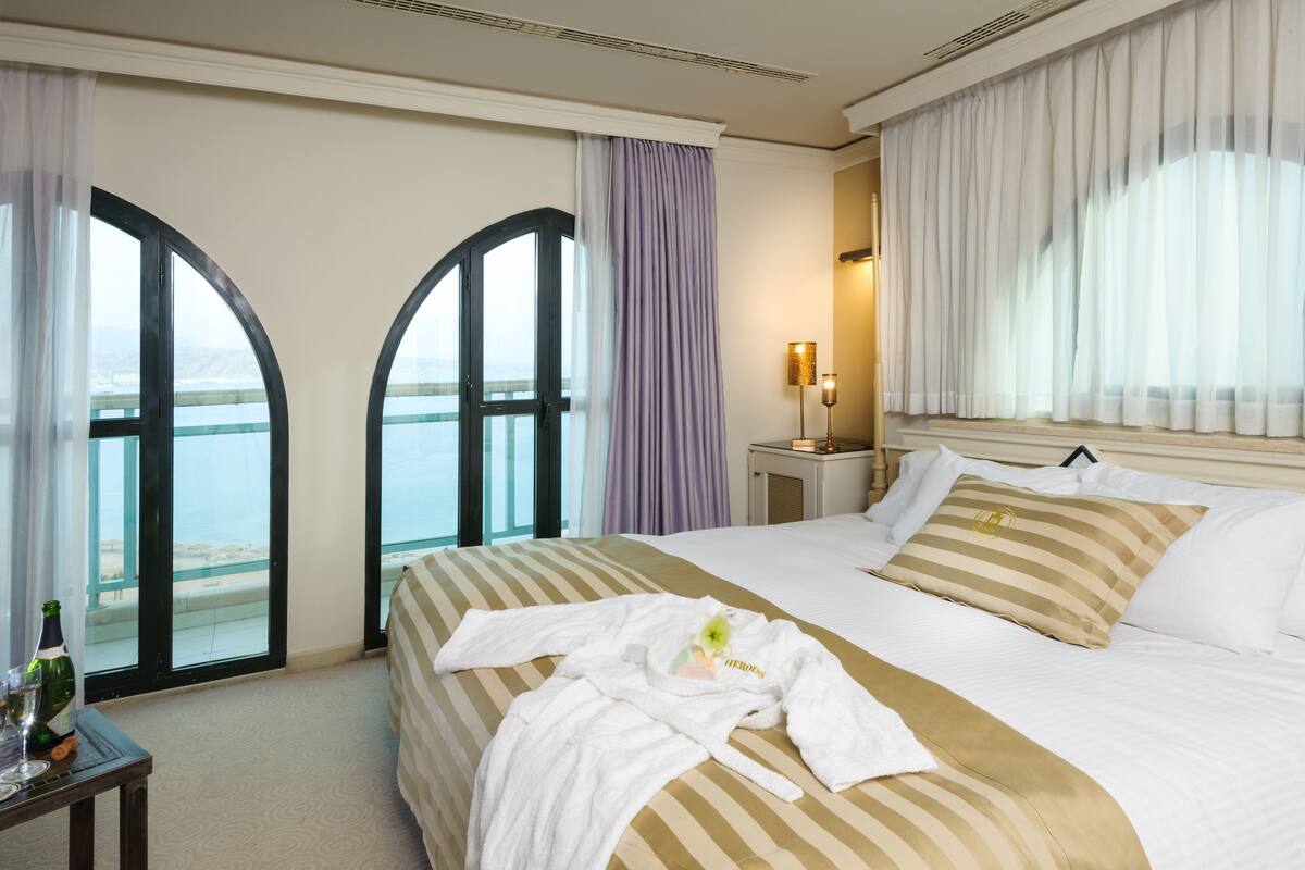 A hotel room with a large bed, a nightstand, a lamp, and a view of the sea through arched windows.