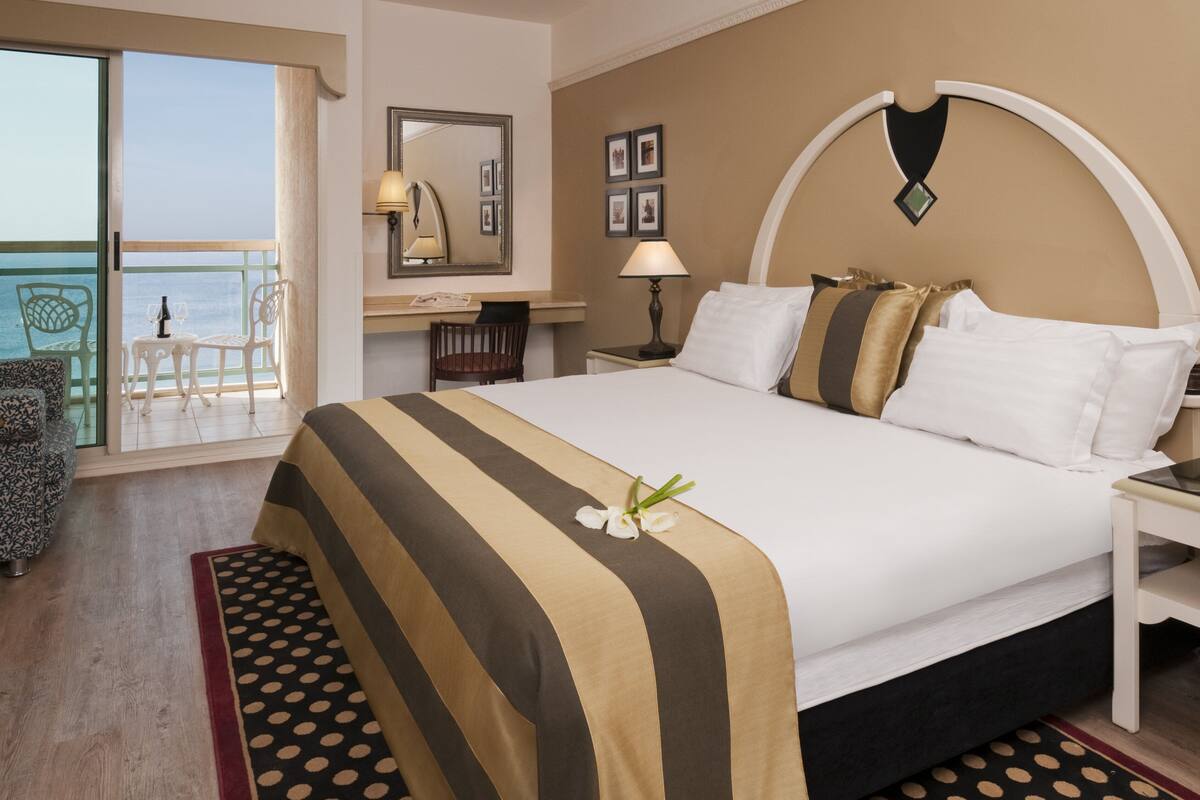 A hotel room with a bed, a desk, a chair, a lamp, and a balcony with a view of the sea.