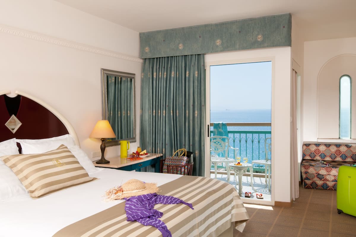 A hotel room with a bed, a balcony with a view, a small table with a lamp, and a chair.
