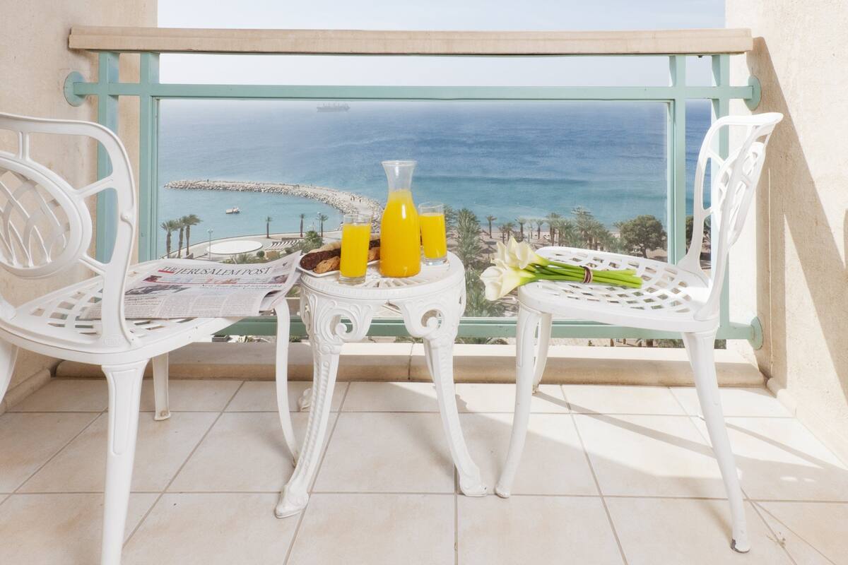 A balcony with two white chairs, a small table with a newspaper, a glass of orange juice, a bottle of orange juice, and a bouquet of flowers.