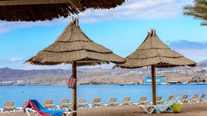 A beach with thatched umbrellas and sun loungers facing a calm sea and distant mountains. - Herods Palace Hotel (Eilat)