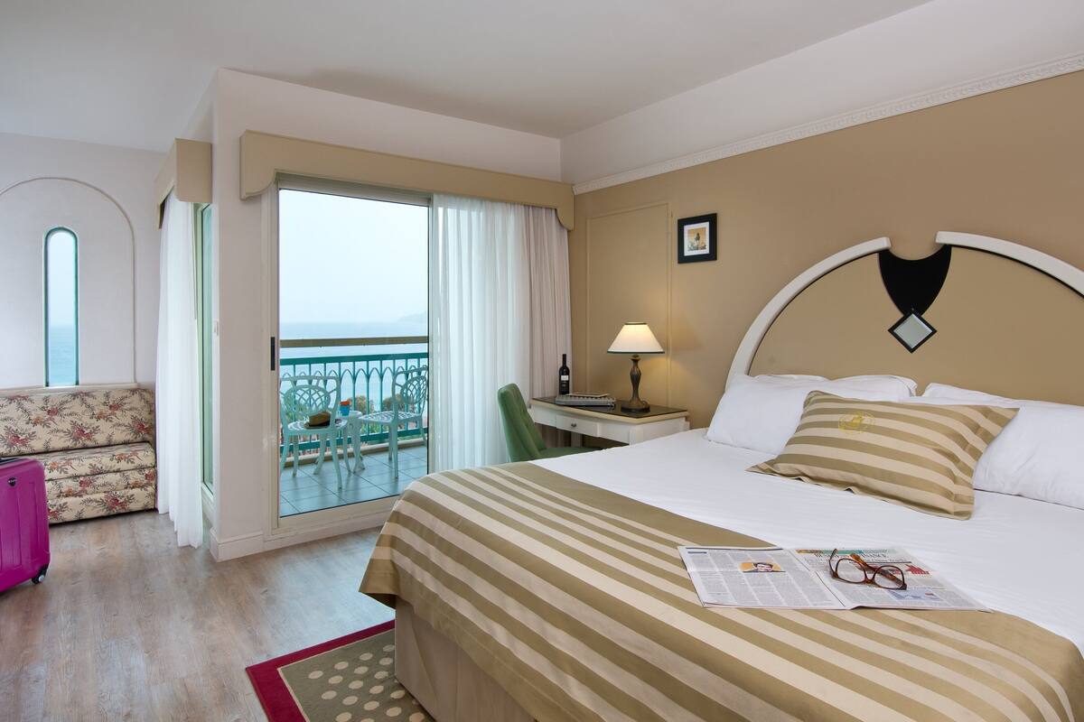 A hotel room with a bed, a desk, a chair, a window with curtains, and a view of the sea.