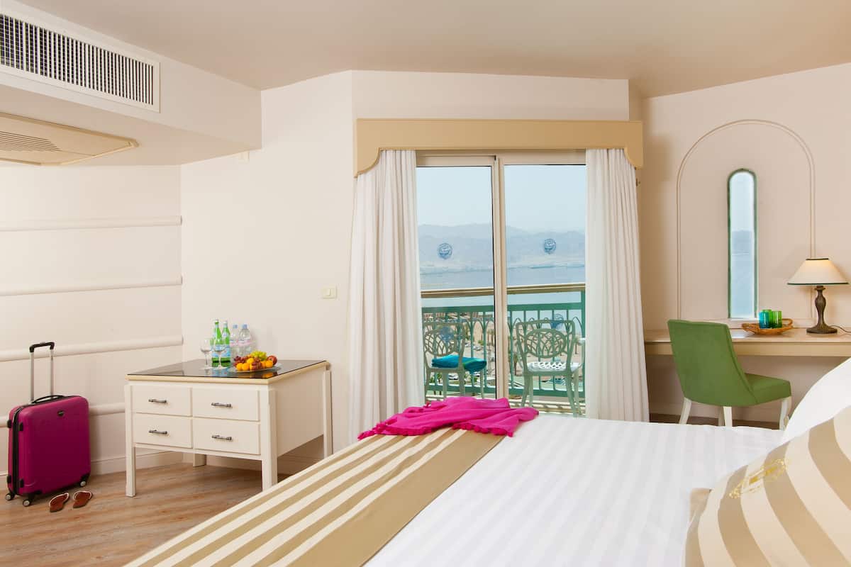 A hotel room with a bed, a desk with a chair, a suitcase, a window with a view, and a balcony with chairs.